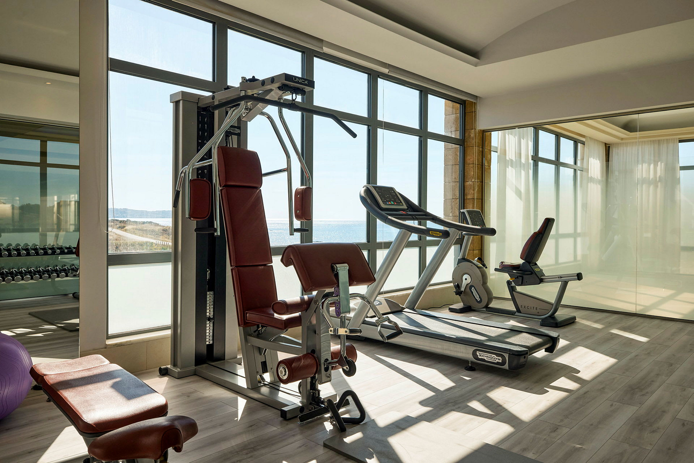 Ett gym med träningsmaskiner, inklusive löpband och styrketräningsutrustning, med stora fönster som ger utsikt mot landskapet utanför. (AI-genererad text)