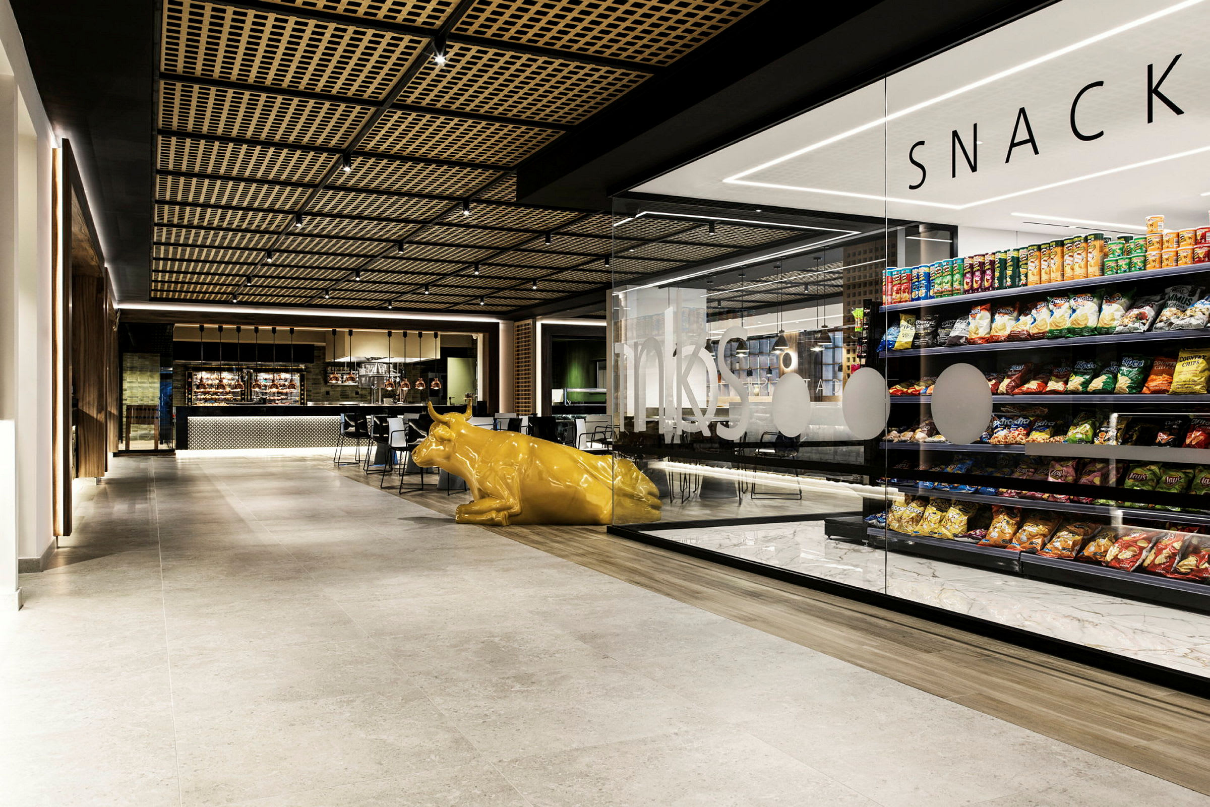 En inomhusbild av ett modernt kafé med butikshyllor fyllda med snacks och en gul kofigur i främre delen. (AI-genererad text)