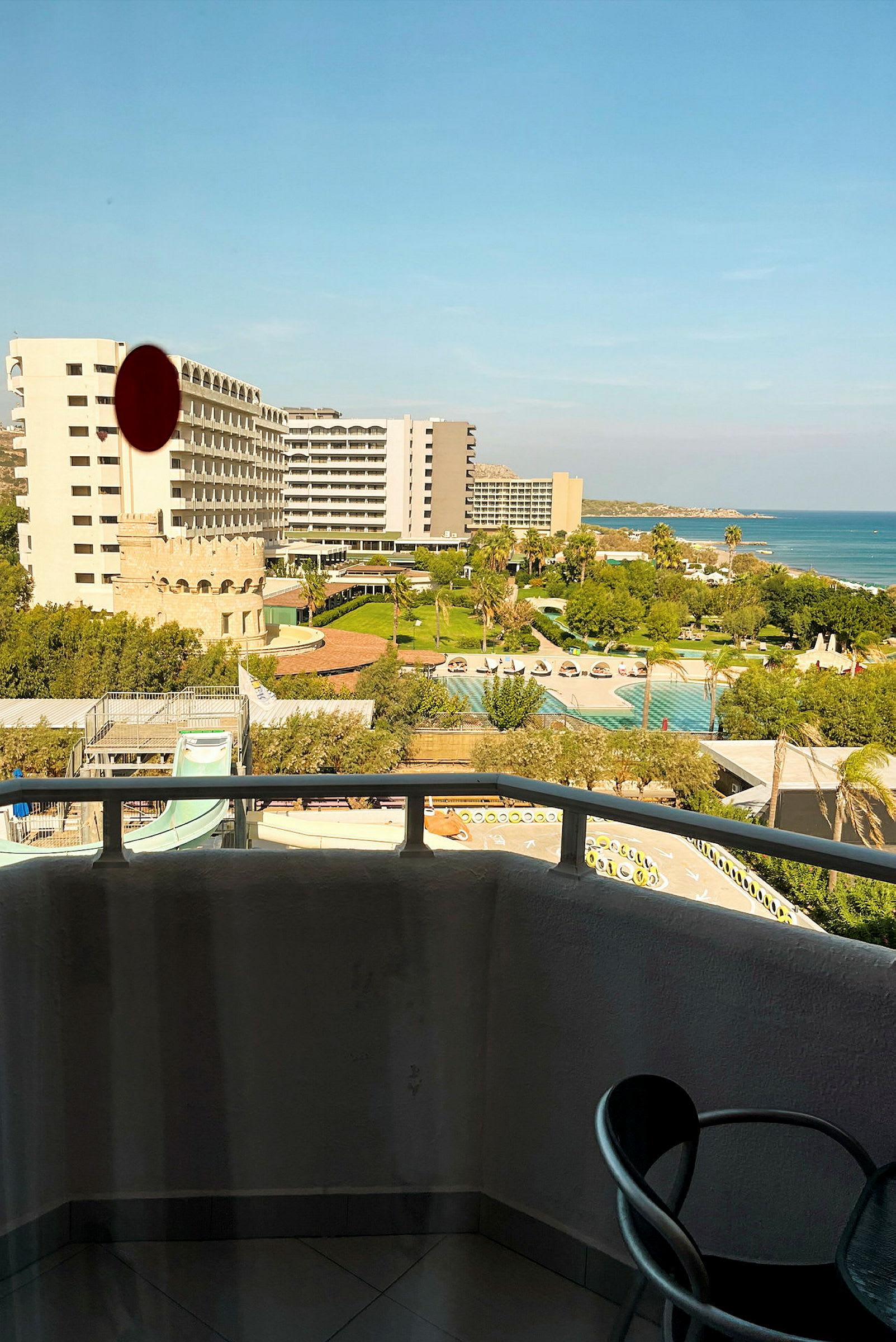 Utsikt från en balkong med en stol och räcke, med hotellbyggnader, poolområde och havet i bakgrunden. (AI-genererad text)