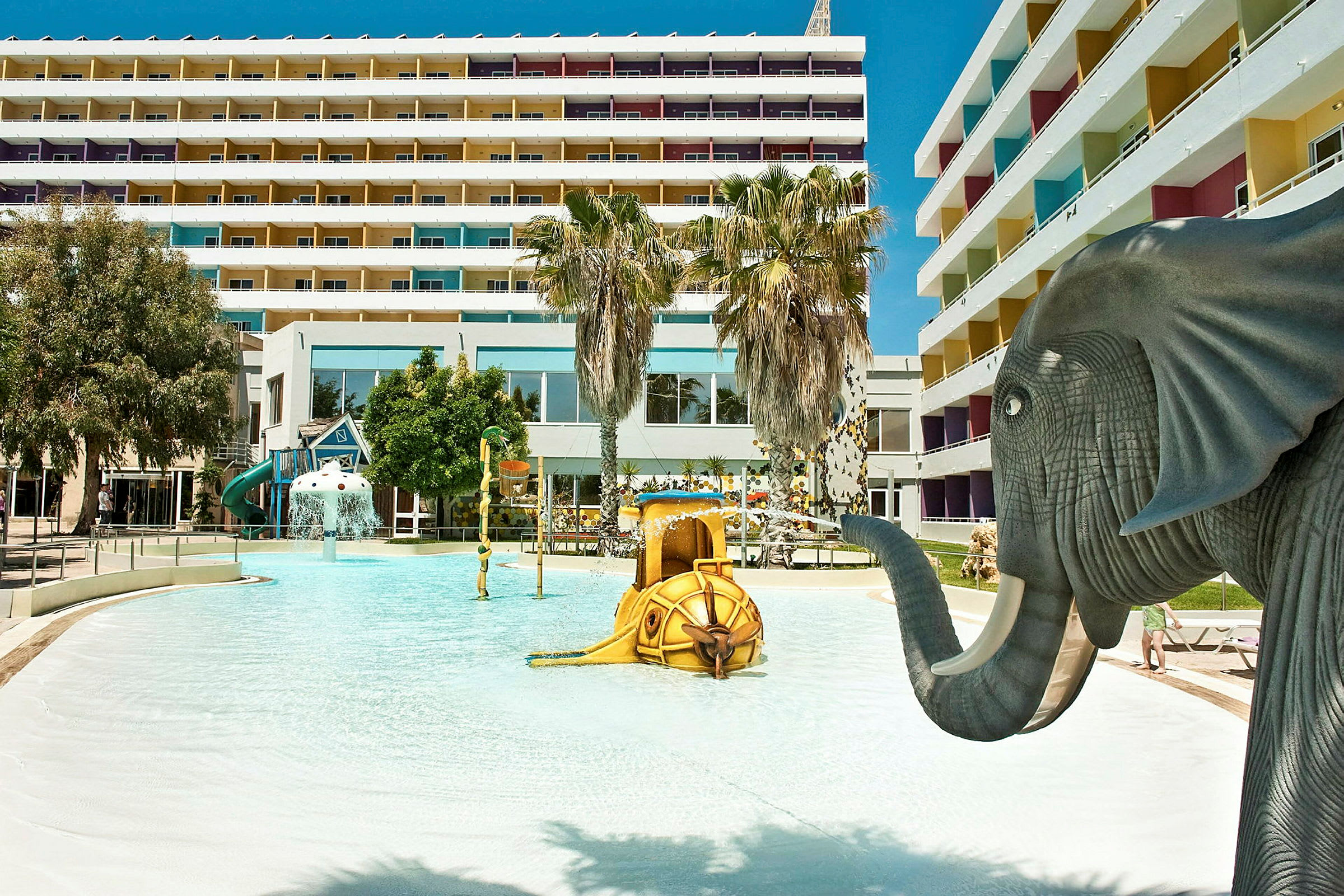Barnpool med elefantfigur och lekskulpturer vid hotellkomplex. (AI-genererad text)