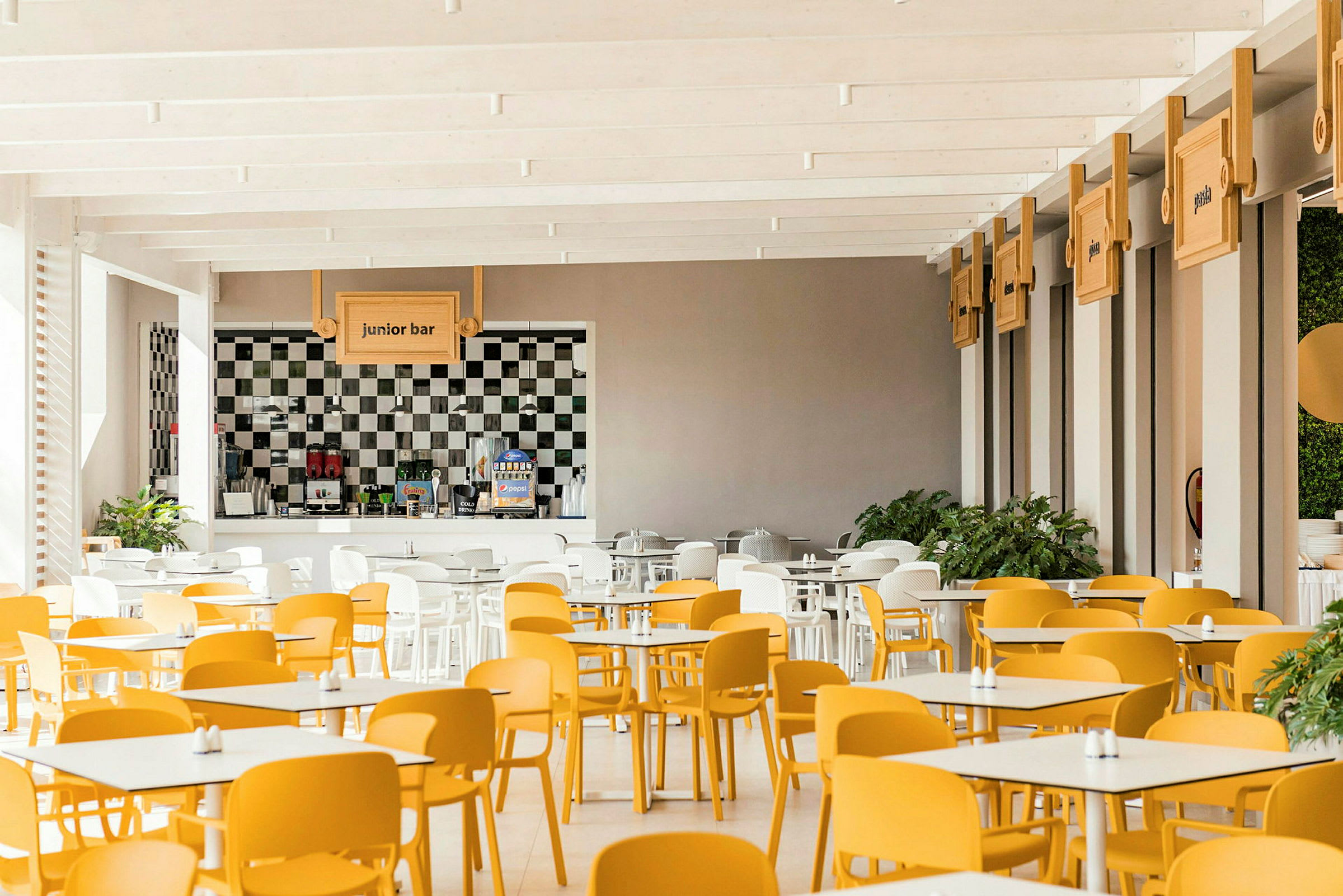 En ljus matsal med gula och vita stolar samt bordsuppsättning. En bardisk märkt "junior bar" är synlig i bakgrunden. (AI-genererad text)