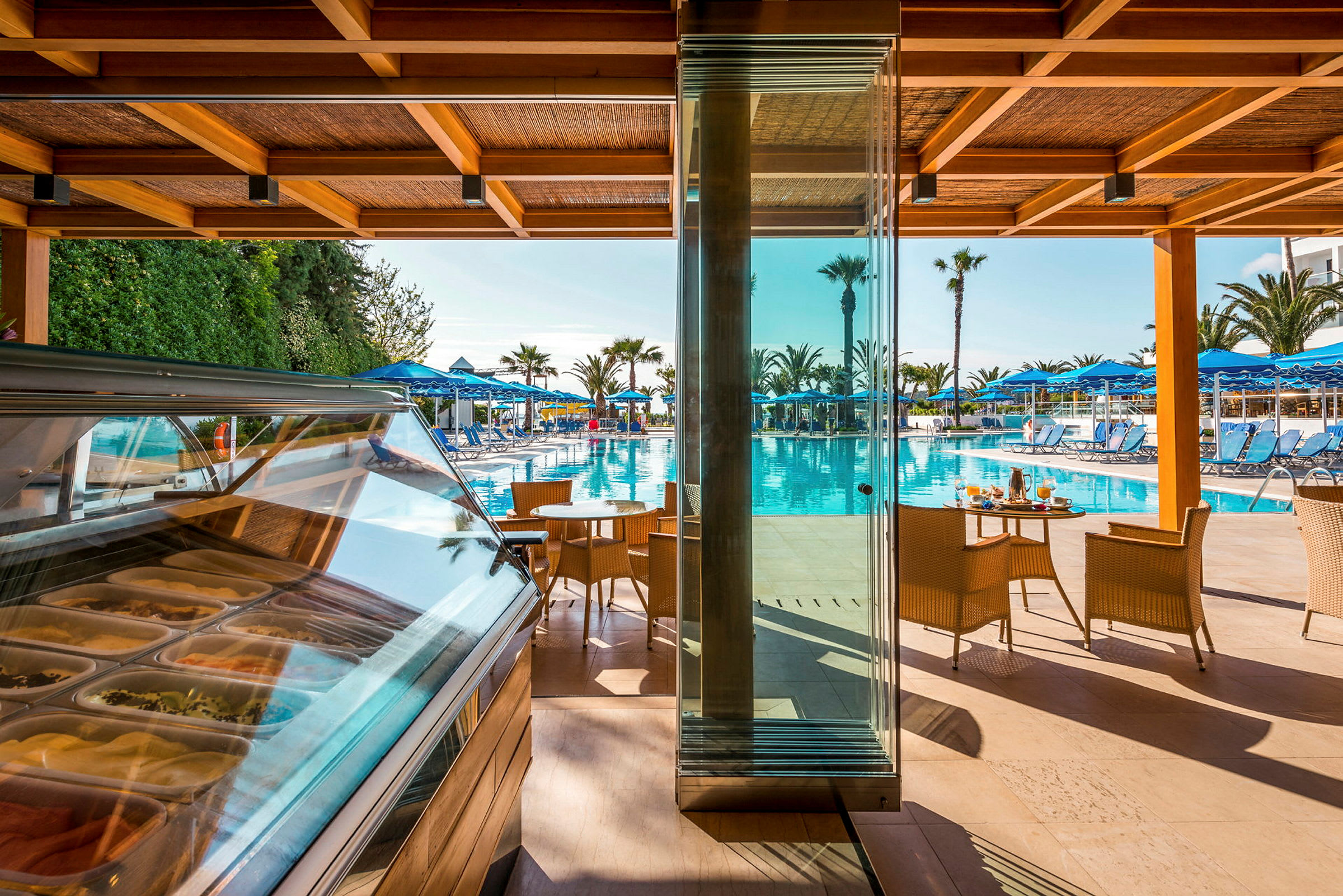 Vy över en pool med solstolar och parasoller sett från en terrass med glasväggar och sittmöbler. (AI-genererad text)