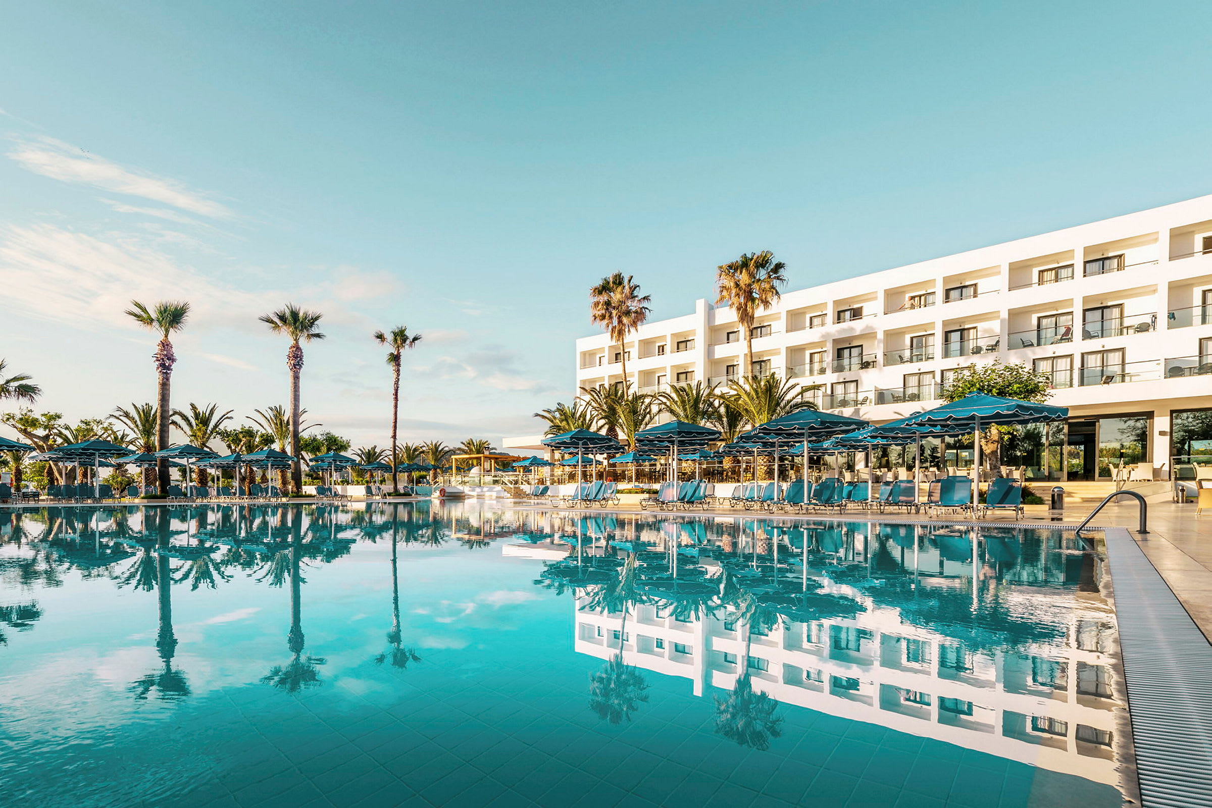 En pool med solstolar och parasoller framför ett modernt hotell med palmträd. (AI-genererad text)