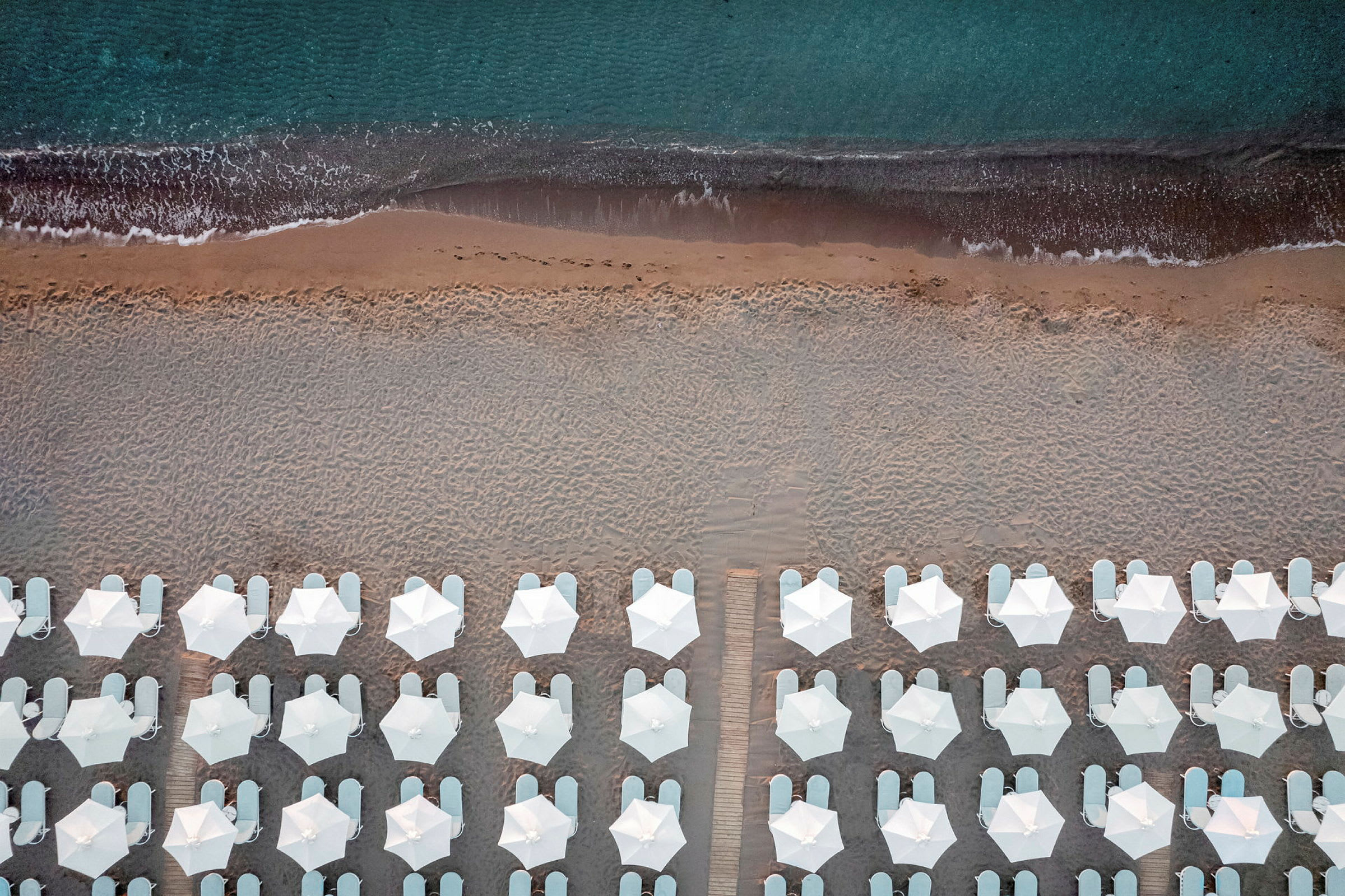 Fågelspråksperspektiv av strand med vita parasoller och solstolar vid kanten av havet. (AI-genererad text)