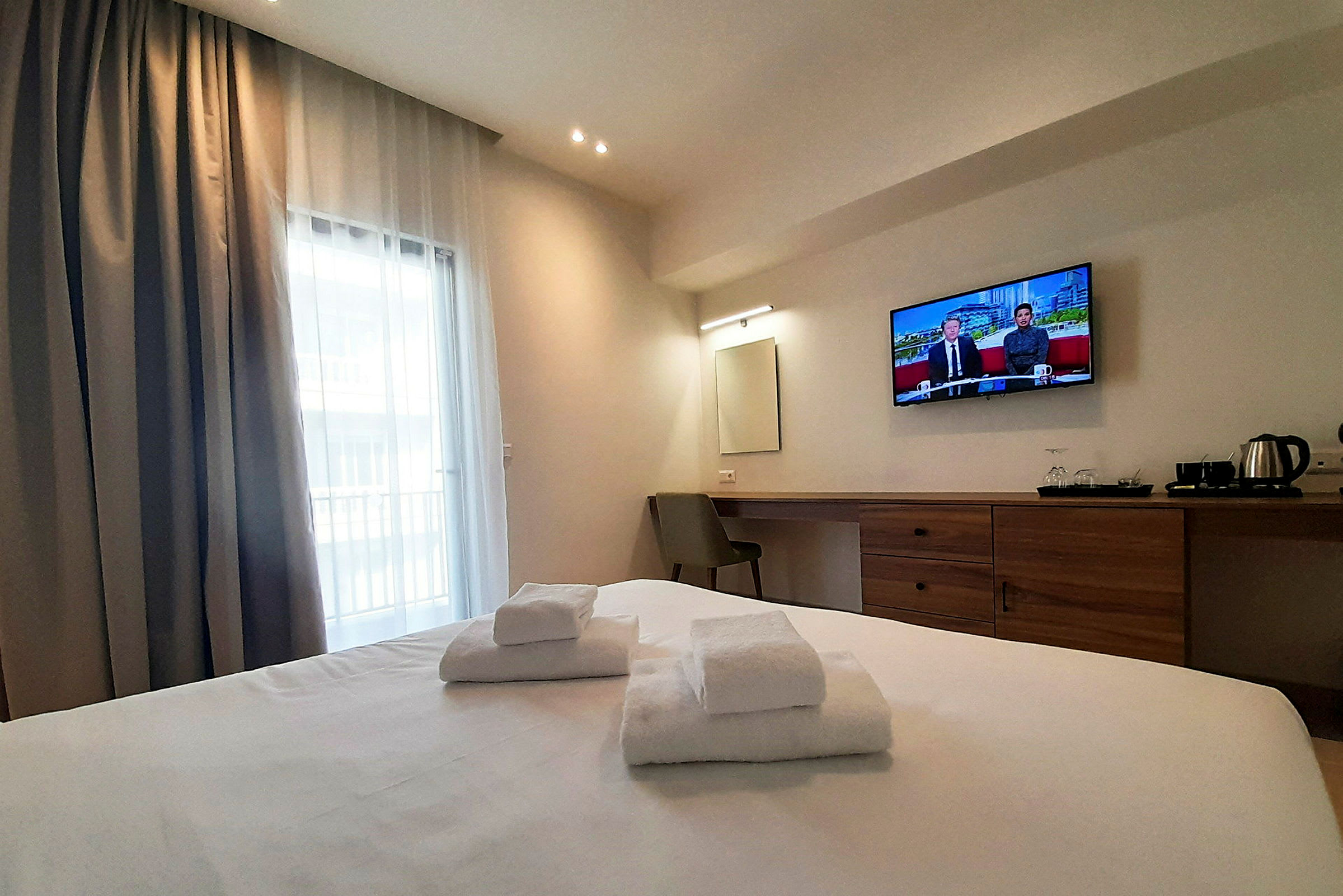 Ett hotellrum med en dubbelsäng, vita handdukar, en väggmonterad tv och ett skrivbord med stol. Ett fönster med gardiner syns. (AI-genererad text)