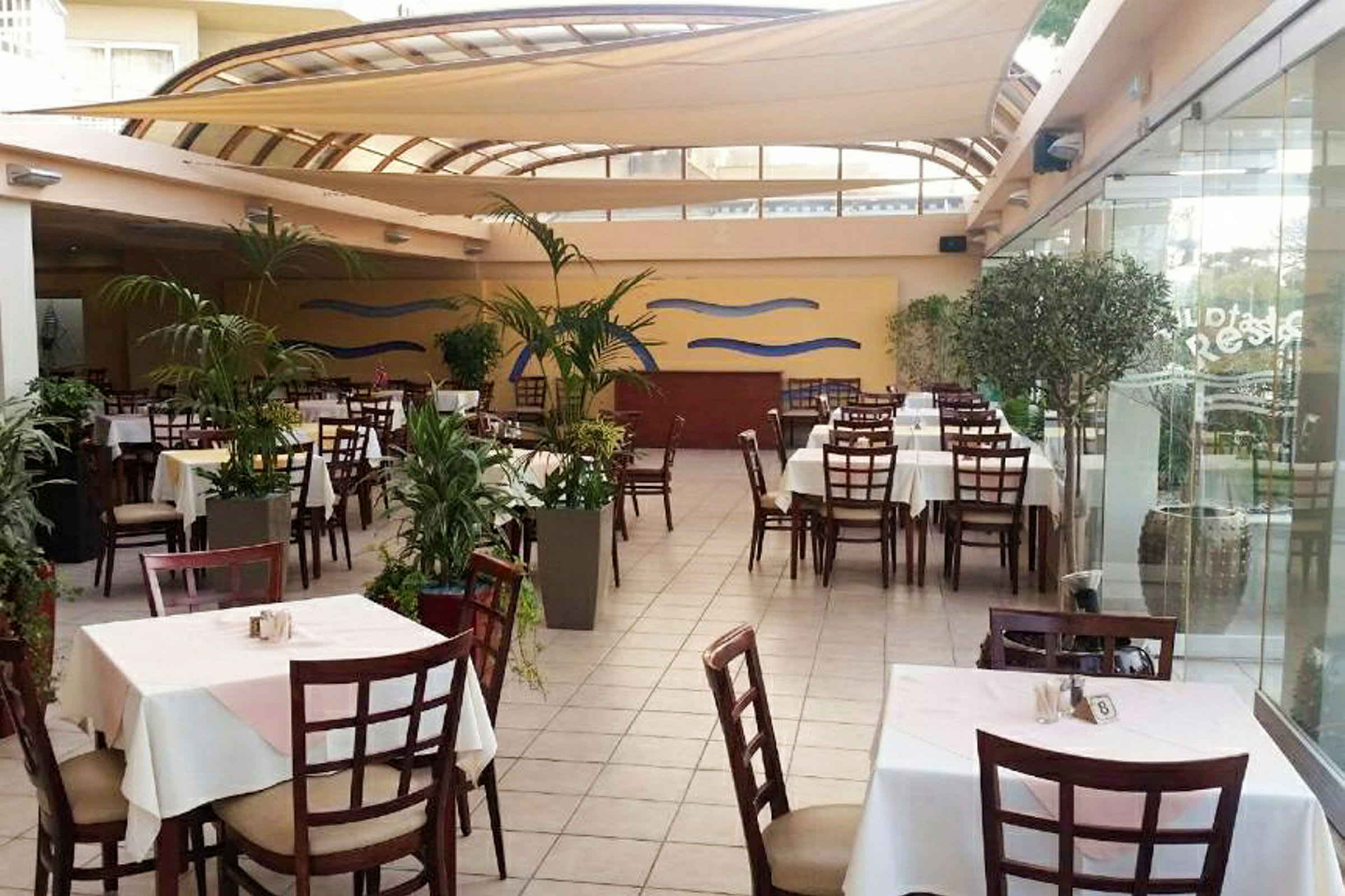 Bild av en restaurang med bord och stolar, omgiven av växter. (AI-genererad text)