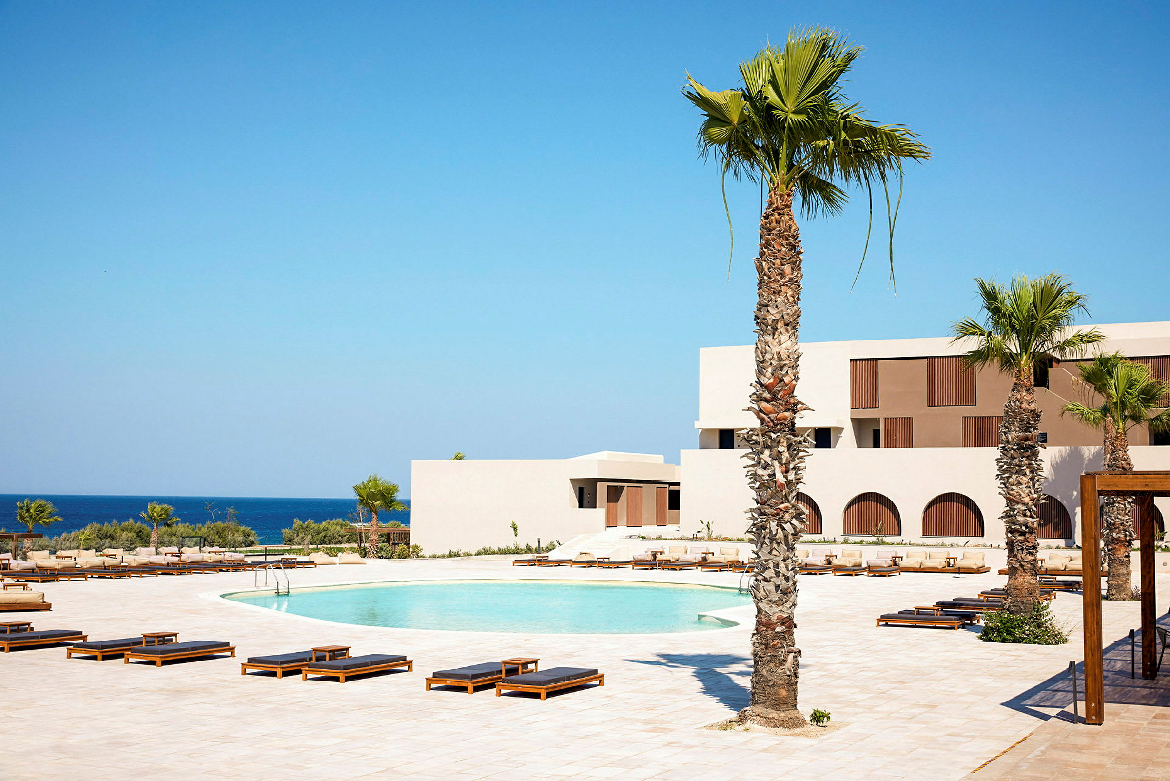Bild av ett hotellområde med palmer, en pool och solstolar framför en vit byggnad nära havet. (AI-genererad text)