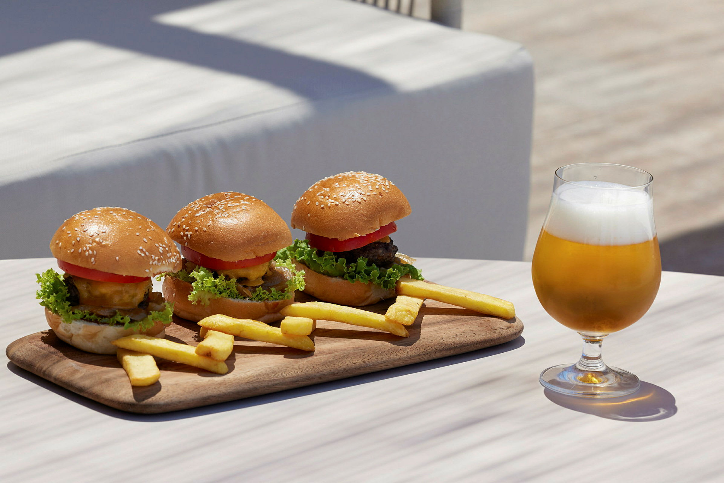 En träbricka med tre hamburgare och pommes frites samt ett glas öl på ett bord. (AI-genererad text)