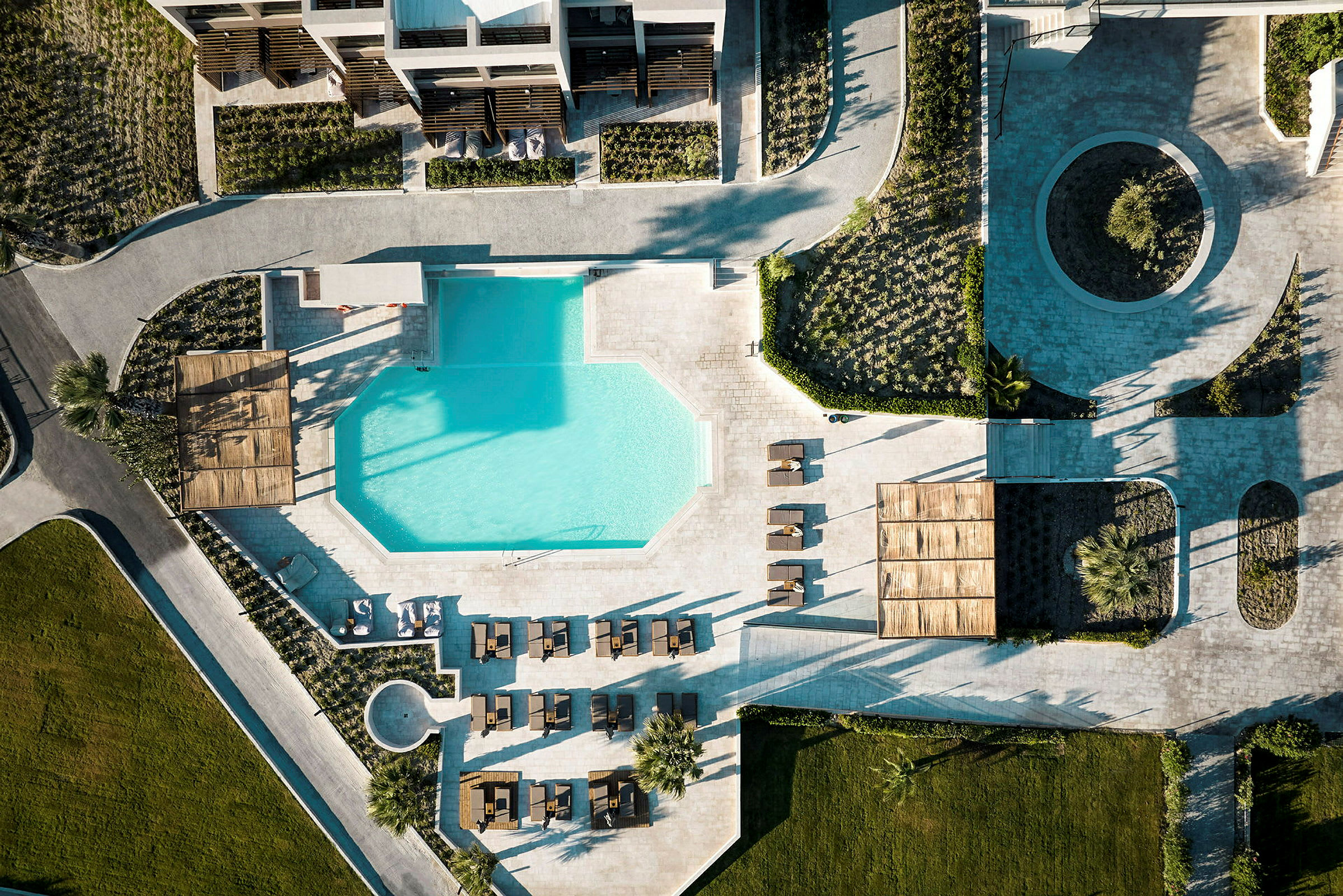 Flygfoto av en resort med en geometriskt formad pool omgiven av solstolar och grönområden. (AI-genererad text)