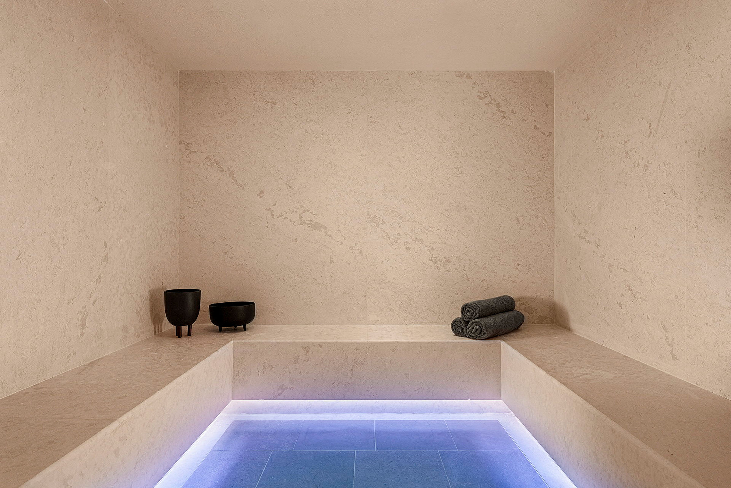 Ett modernt spa-rum med minimalistisk inredning, bänksittplats, grå rullade handdukar och två mörka dekorativa skålar. (AI-genererad text)