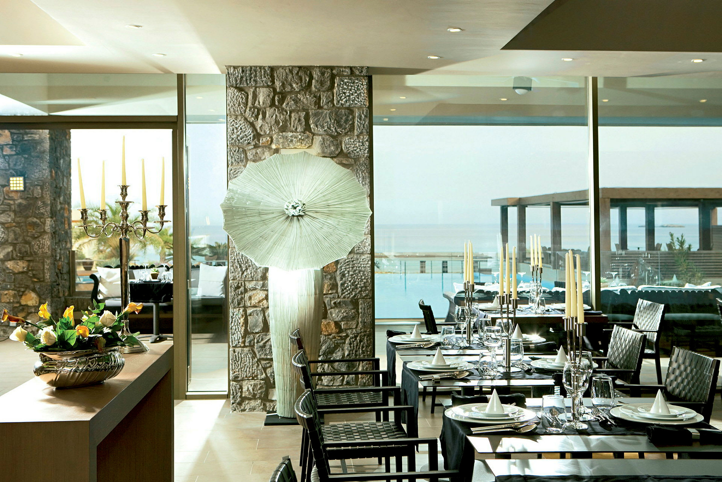 En inomhusrestaurang med dukade bord, en stor fönstervägg med utsikt över en pool och havet samt en dekorerad ljusstake och blommor på ett bord. (AI-genererad text)