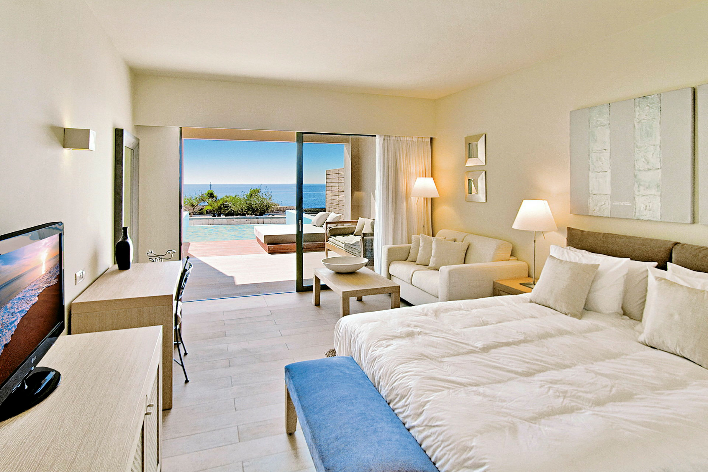 Hotellrum med modern inredning, stor säng, soffa och utsikt mot terrass med pool och hav. (AI-genererad text)
