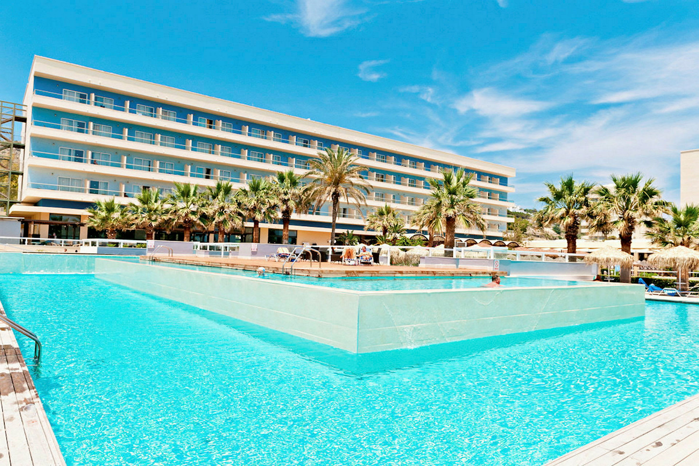 Ett hotell med flera våningar bakom en stor pool omgiven av palmer. (AI-genererad text)