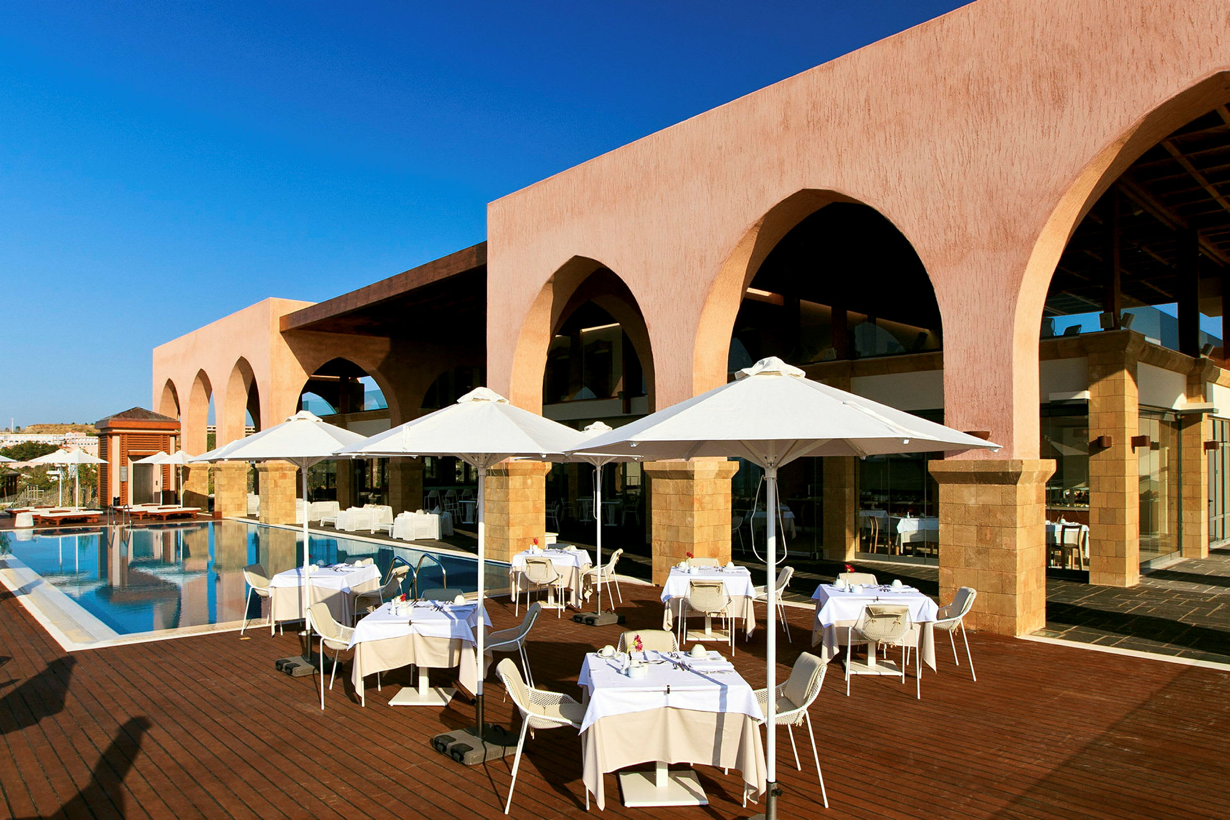 En utomhusrestaurang med vita parasoller vid en pool framför en byggnad med valv. (AI-genererad text)
