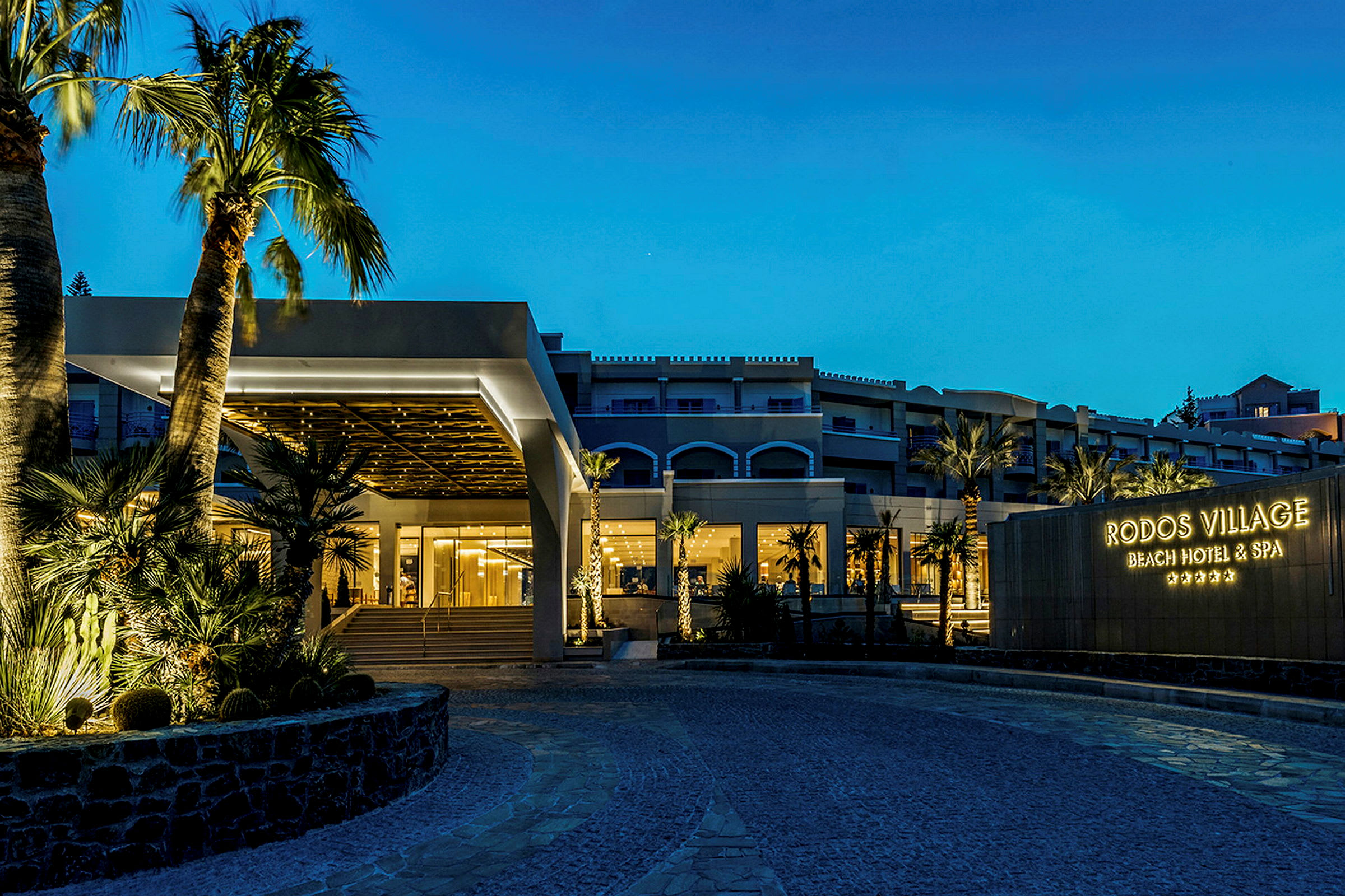 Ett hotell på kvällen med upplyst entré och skylt med texten 'Rodos Village Beach Hotel & Spa'. (AI-genererad text)