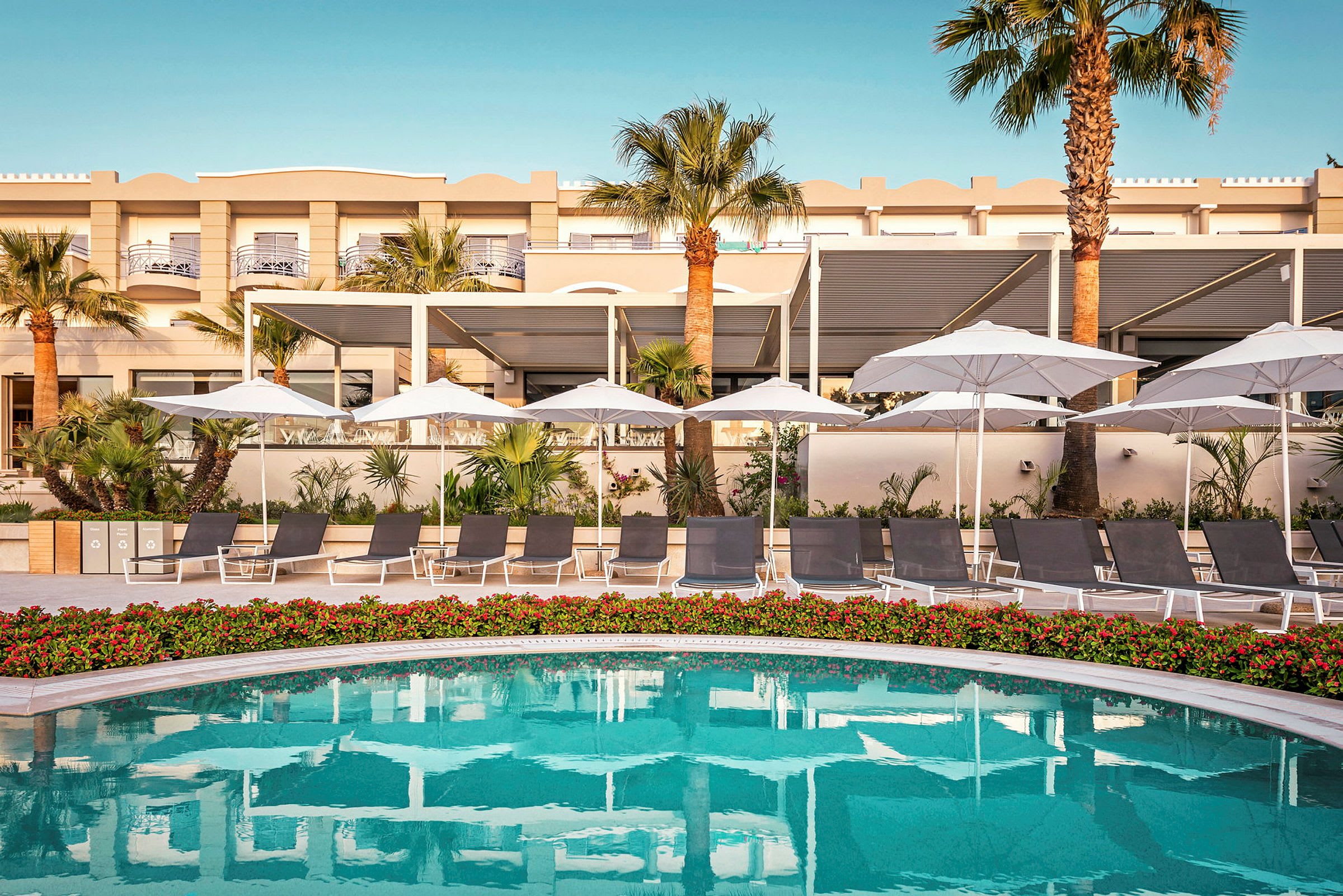 En hotellpool med solstolar och parasoller, omgiven av palmträd och grönska framför själva byggnaden. (AI-genererad text)