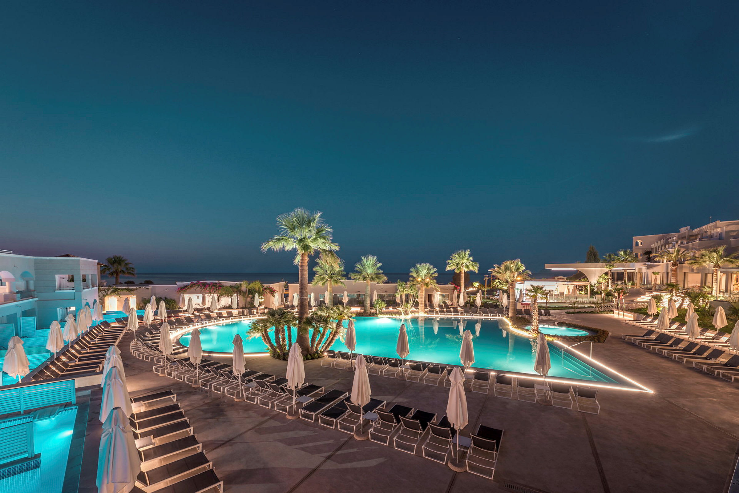 Bild av en upplyst hotellpoolområde med solstolar, parasoller och palmträd i kvällsbelysning. (AI-genererad text)