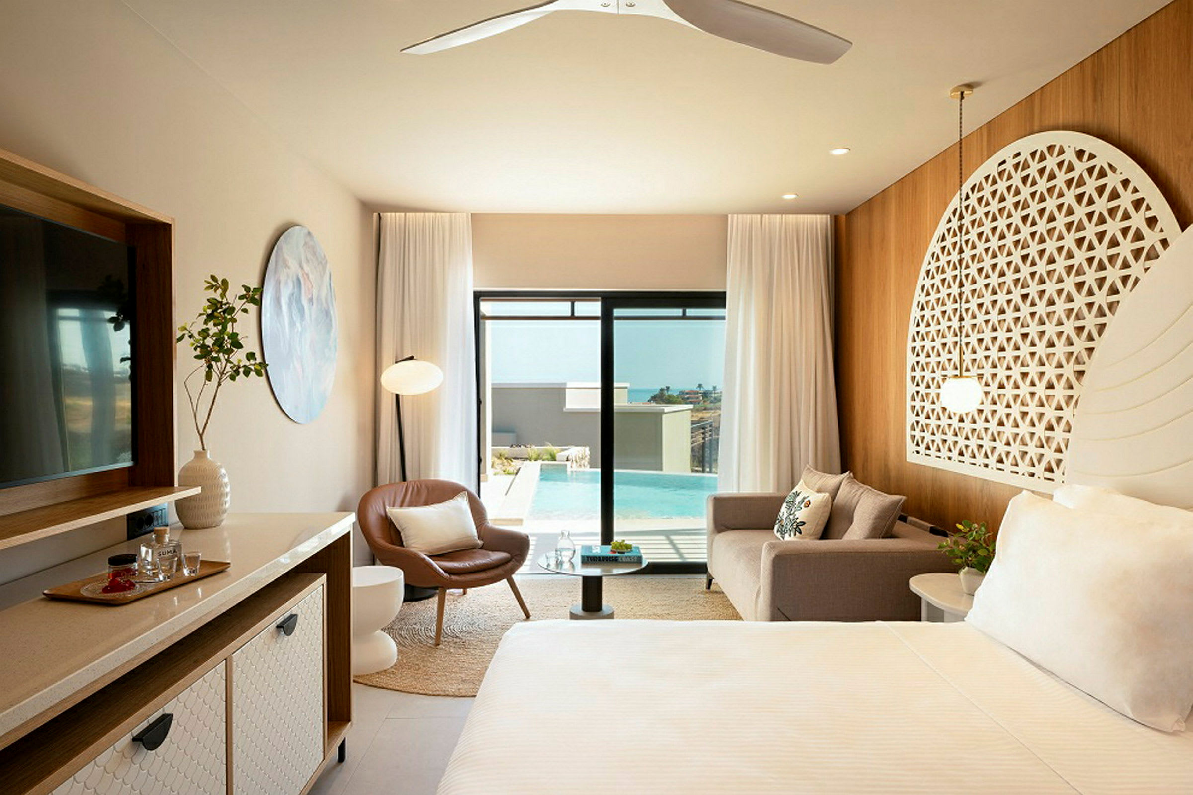 Ett elegant hotellrum med moderna möbler, en soffa, en fåtölj, ett sängområde med prydnadshuvudgavel, en stor spegel och glasdörrar som öppnar mot en swimmingpool utomhus. (AI-genererad text)