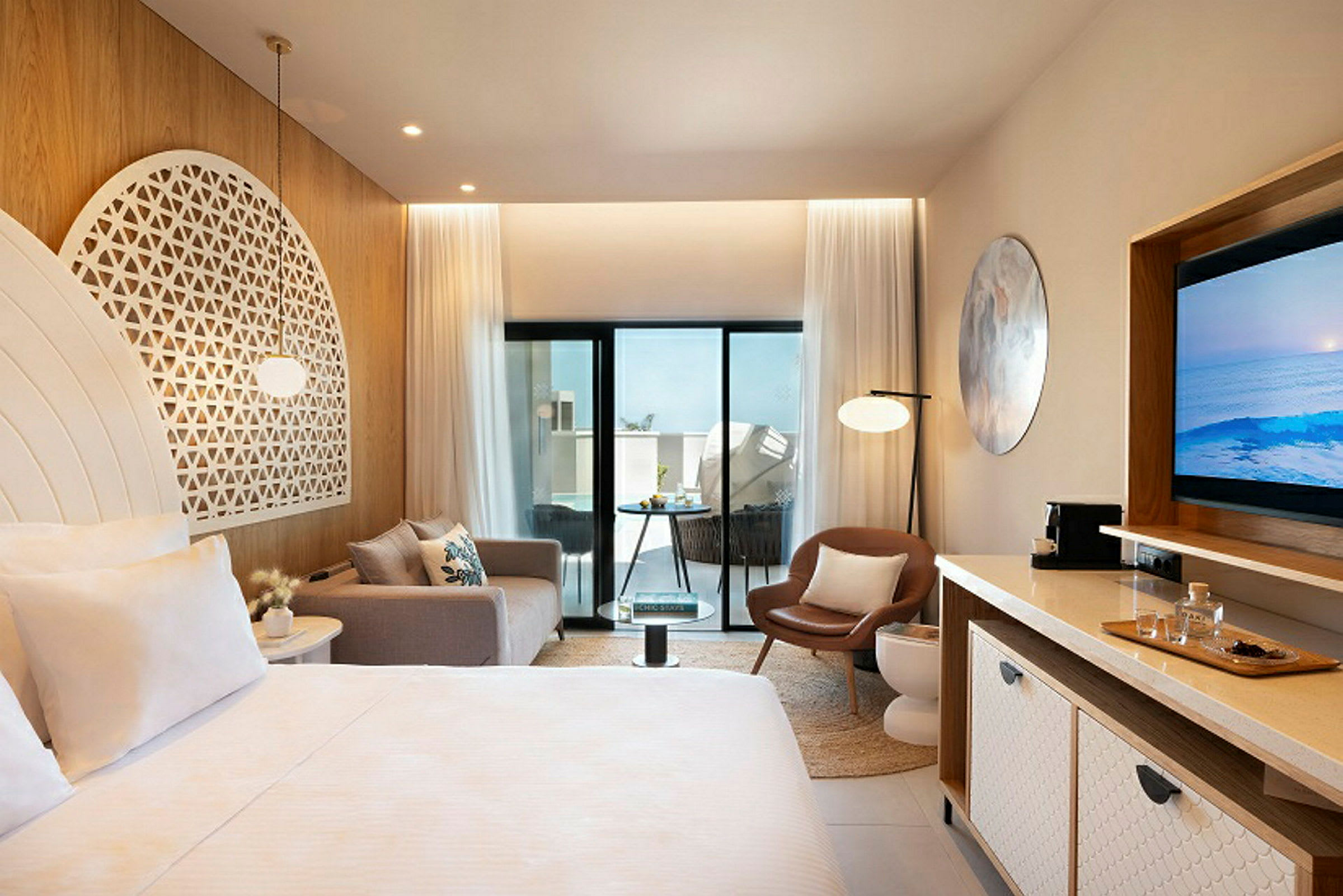 Ett modernt hotellrum med ljusa möbler och dekoration, inkluderande en säng, soffa, stol och TV. Rummet har stora glasdörrar som leder ut till en balkong. (AI-genererad text)