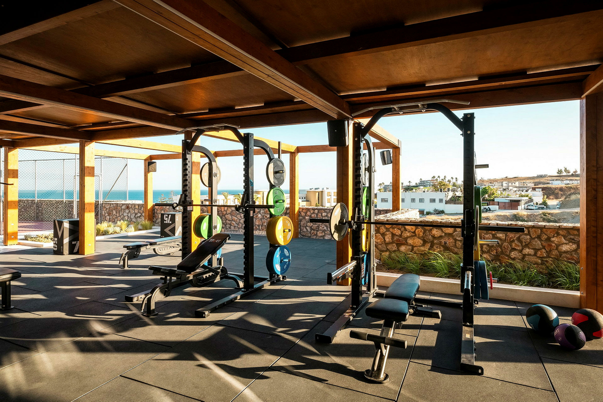 Ett utomhusgym med träningsutrustning, inklusive vikter och bänkar, under ett tak med utsikt över ett landskap och havet. (AI-genererad text)