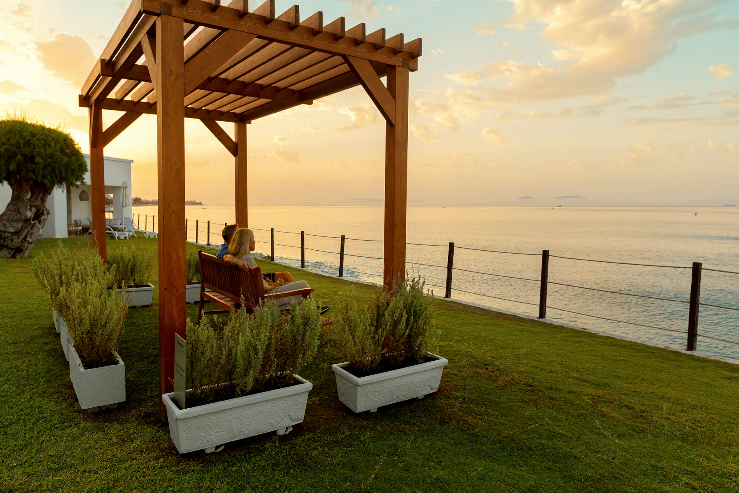 Personer sitter under träpergola med utsikt över havet. (AI-genererad text)