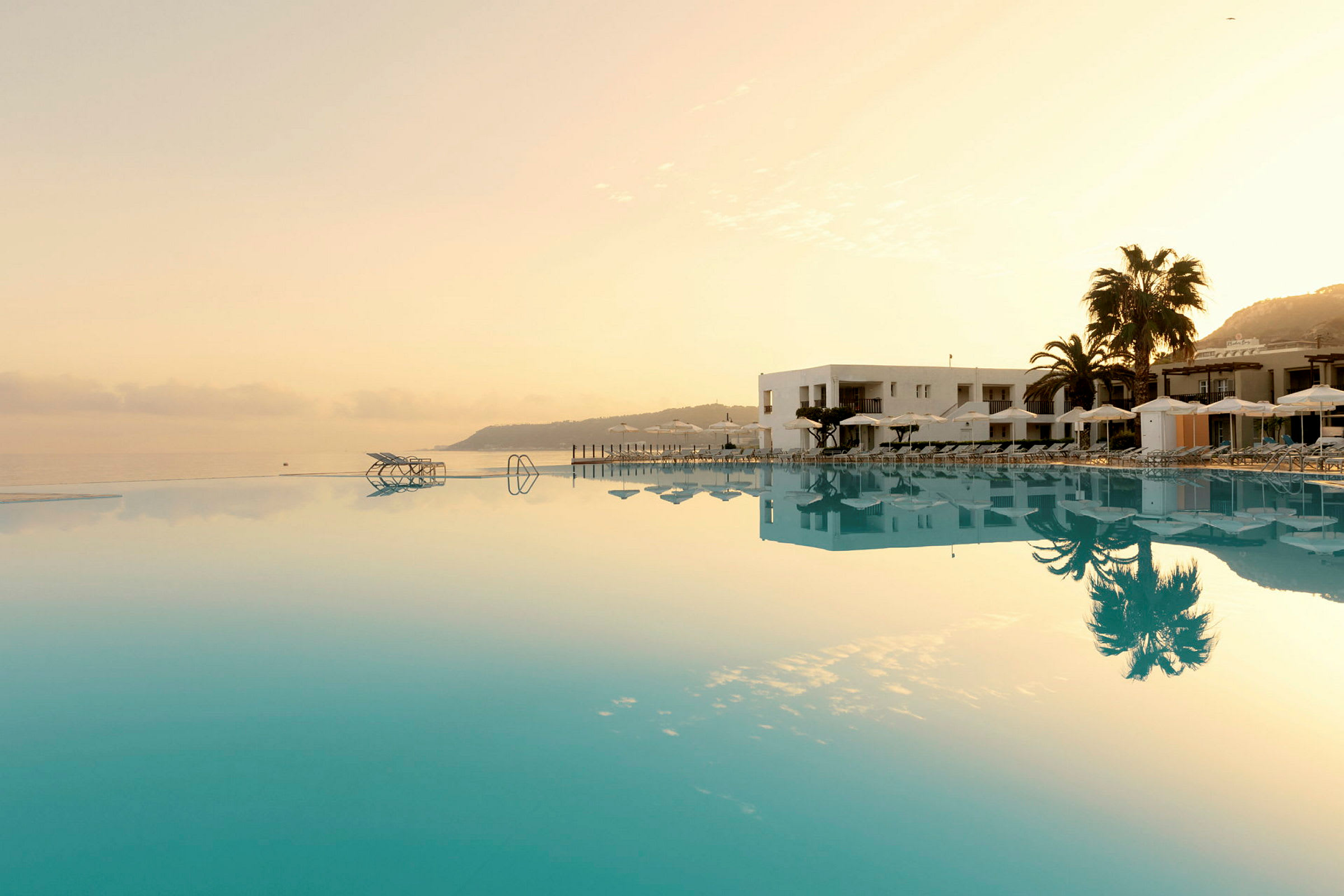 Bilden visar en infinitypool vid sidan av ett hotell med solstolar och parasoller, omgiven av palmer och morgonljus. (AI-genererad text)