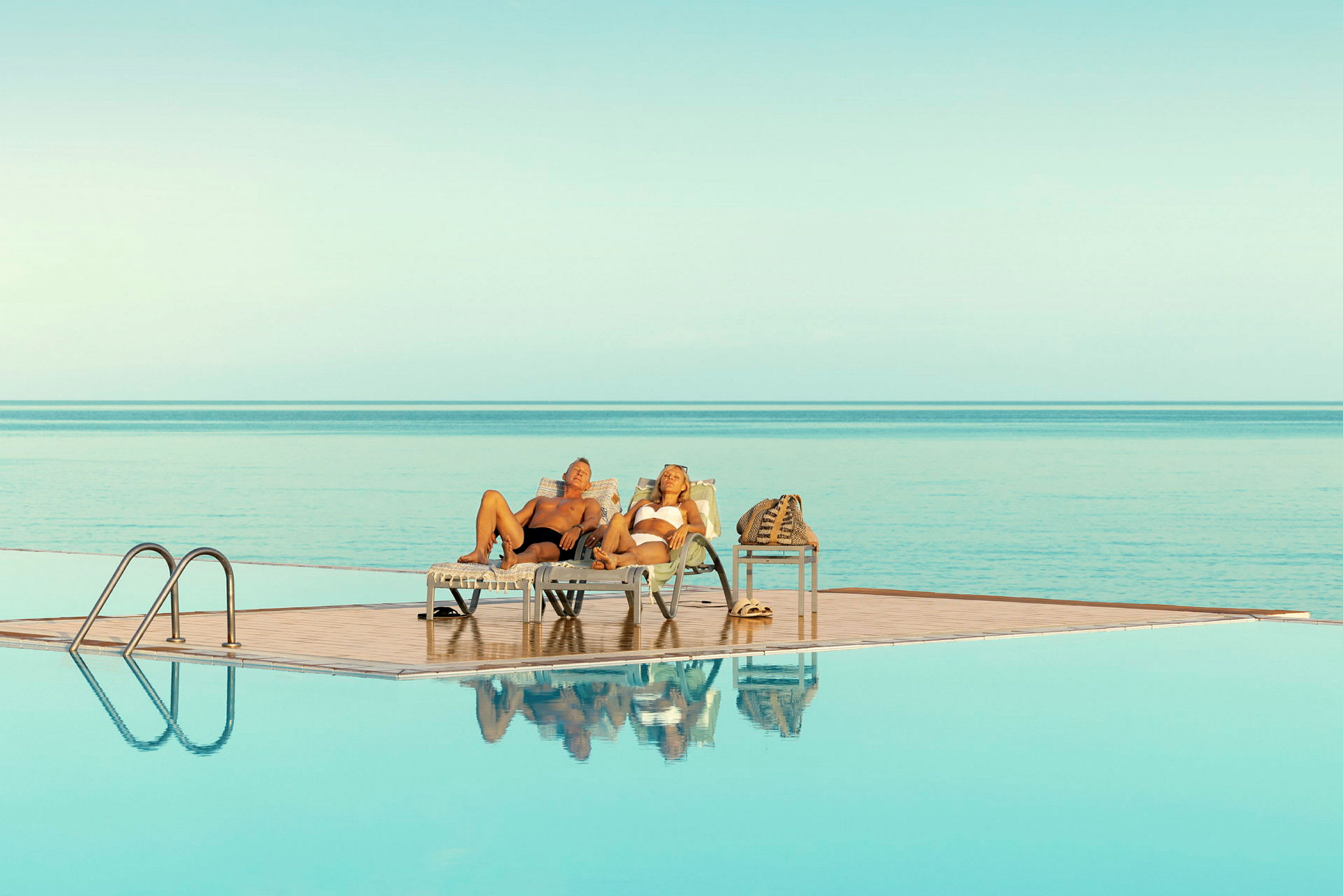 Två personer ligger på solstolar vid en pool med utsikt över havet. (AI-genererad text)