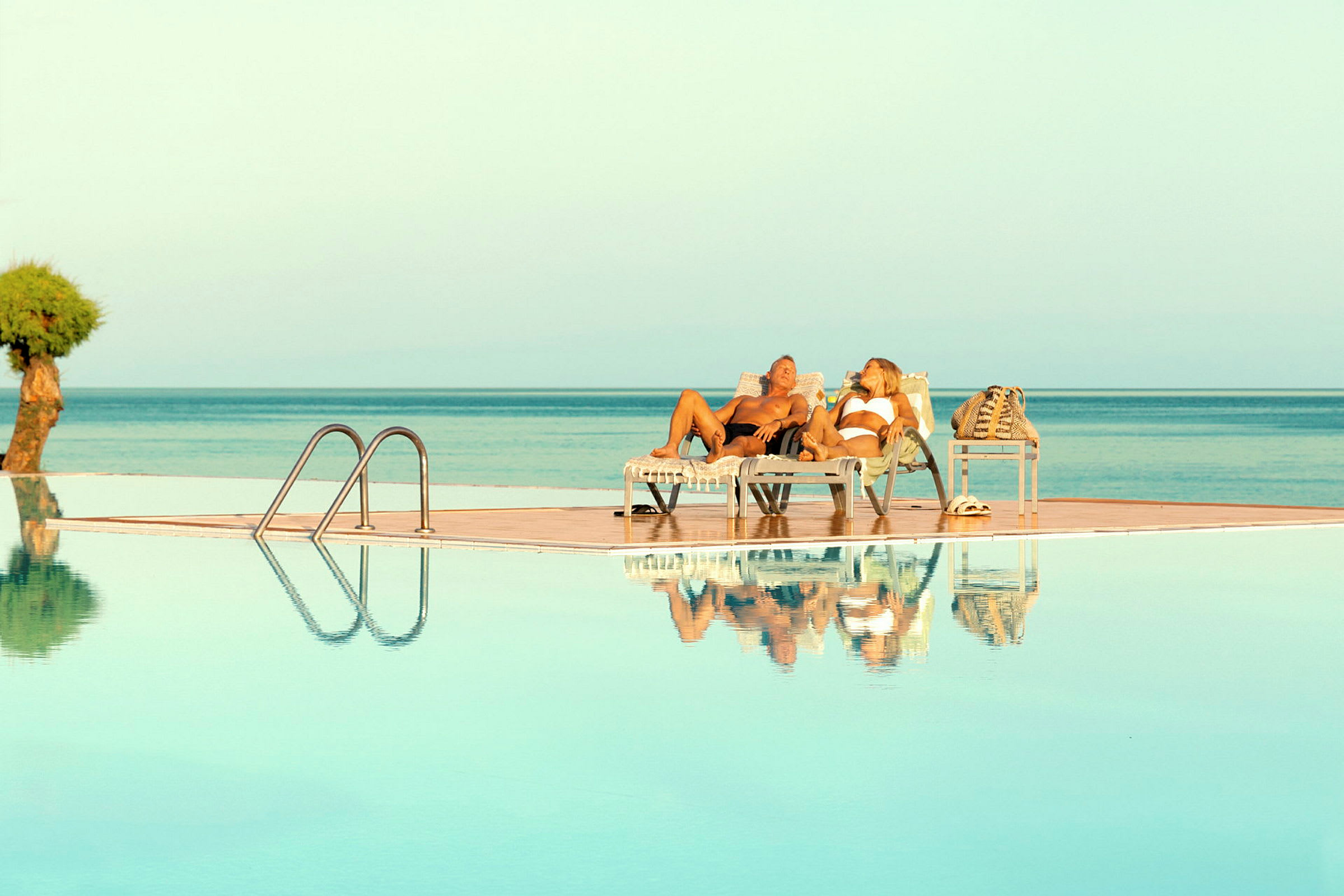Två personer ligger på solstolar vid en pool med utsikt över havet. (AI-genererad text)
