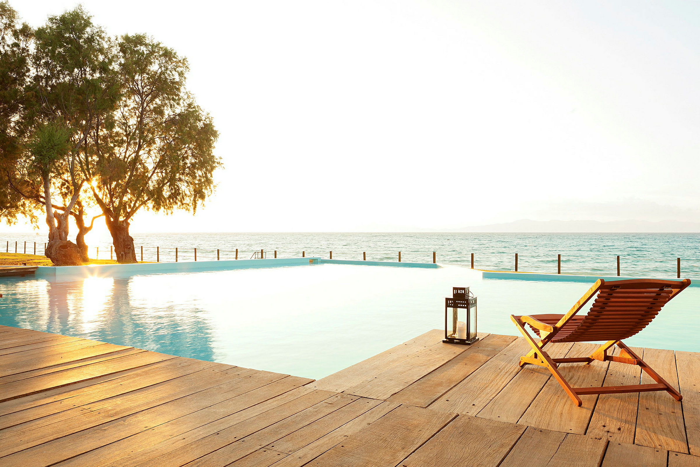 Fotografi av en infinitypool mötandes havet vid solnedgång, med en trästol och en lykta på en träterrass. (AI-genererad text)
