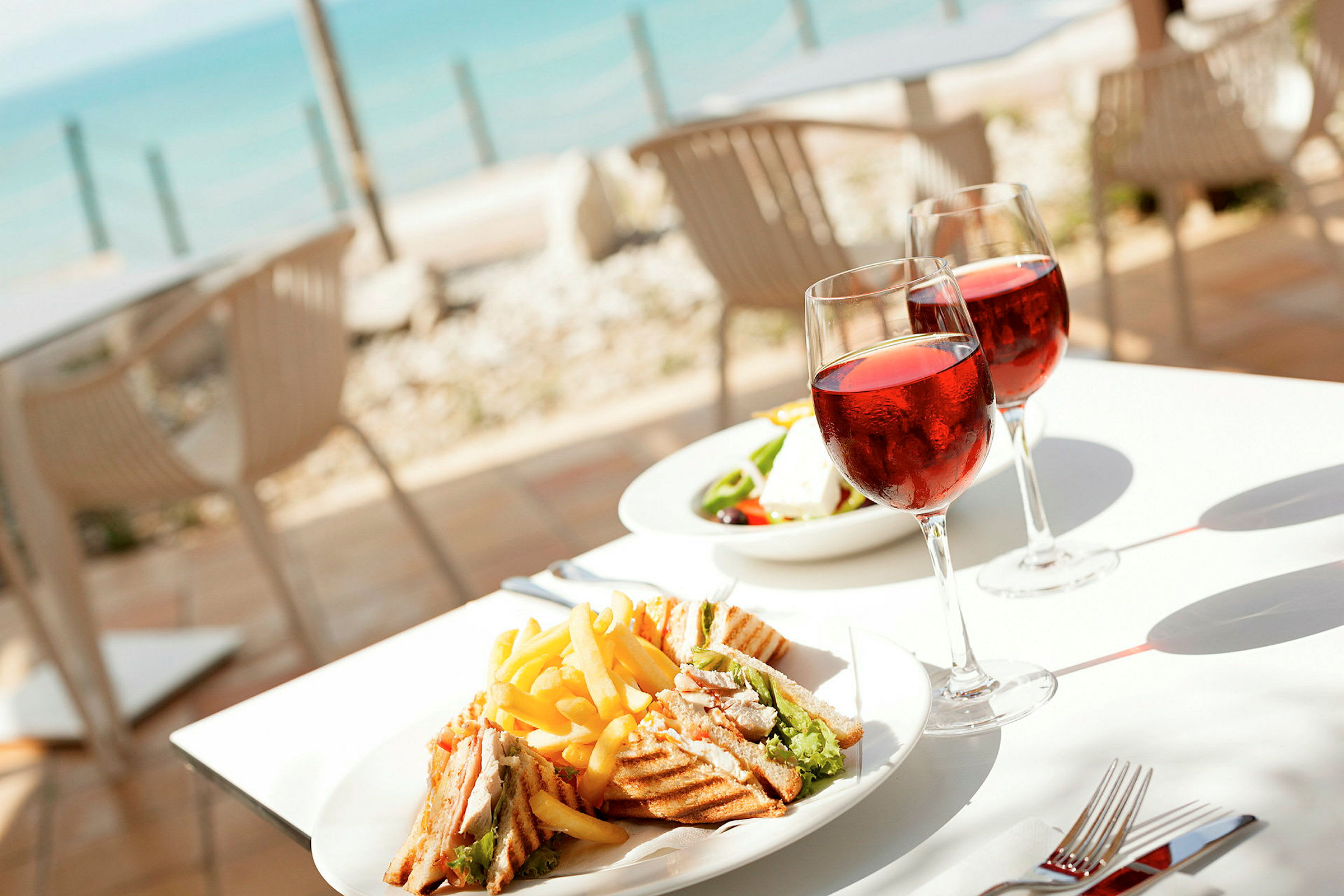 Tallrik med smörgås och pommes frites samt två glas vin vid ett bord nära havet. (AI-genererad text)
