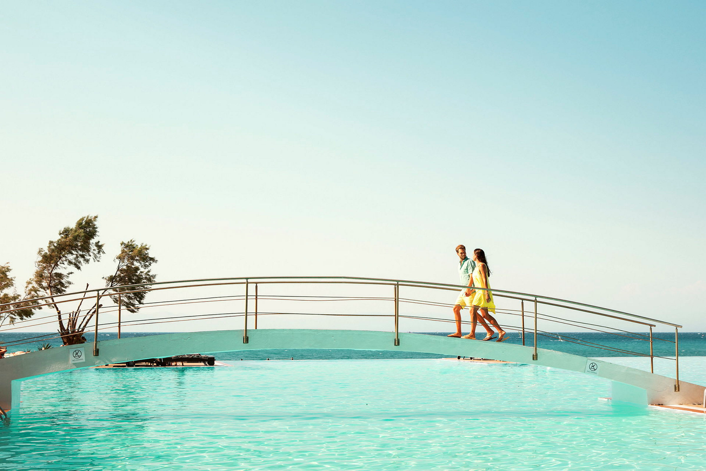 Två personer går över en bro ovanför en pool med havet i bakgrunden. (AI-genererad text)