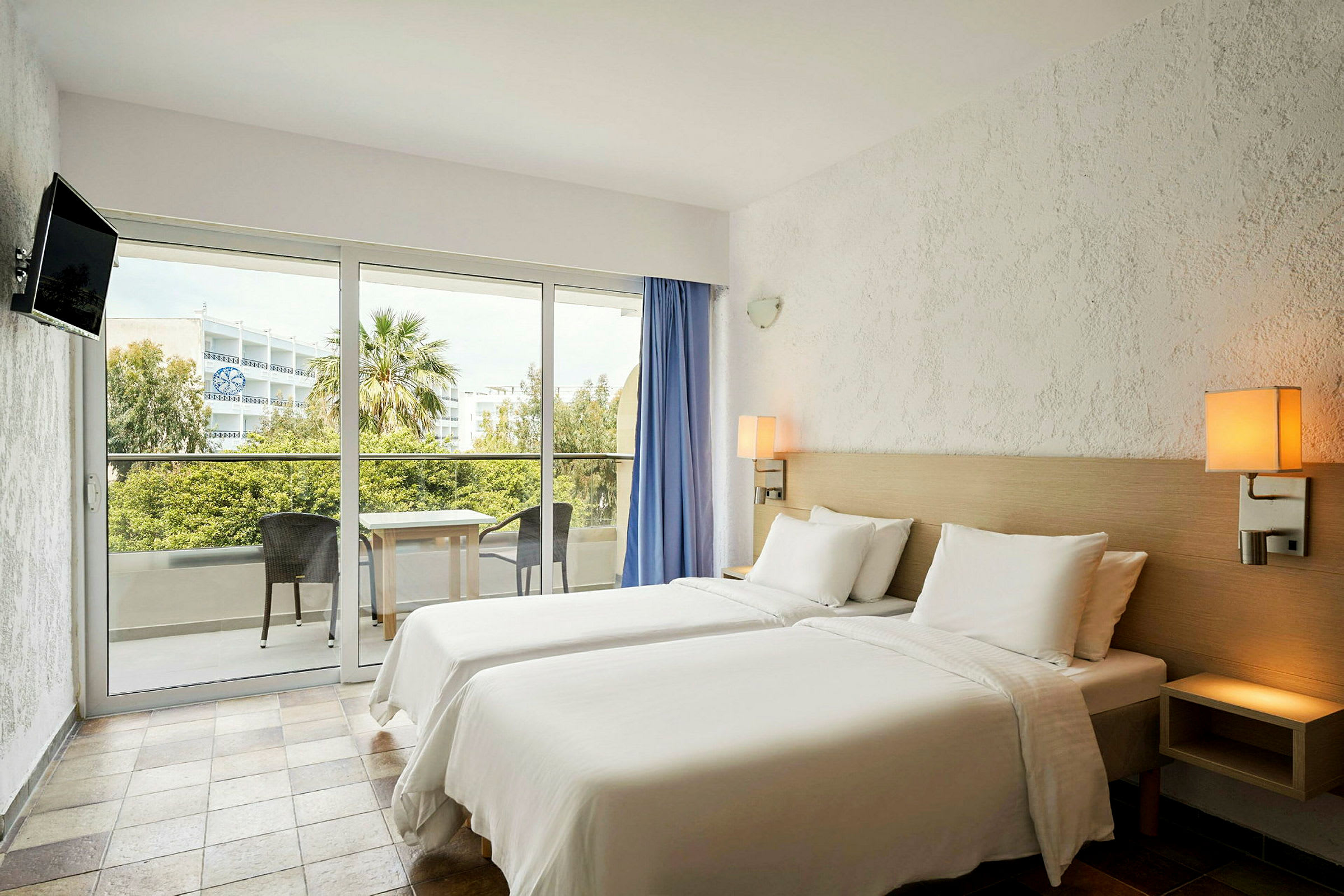 Ett hotellrum med vita sängkläder och två separata sängar, en balkong med utsikt över trädgårdsområdet och en stad i bakgrunden. (AI-genererad text)