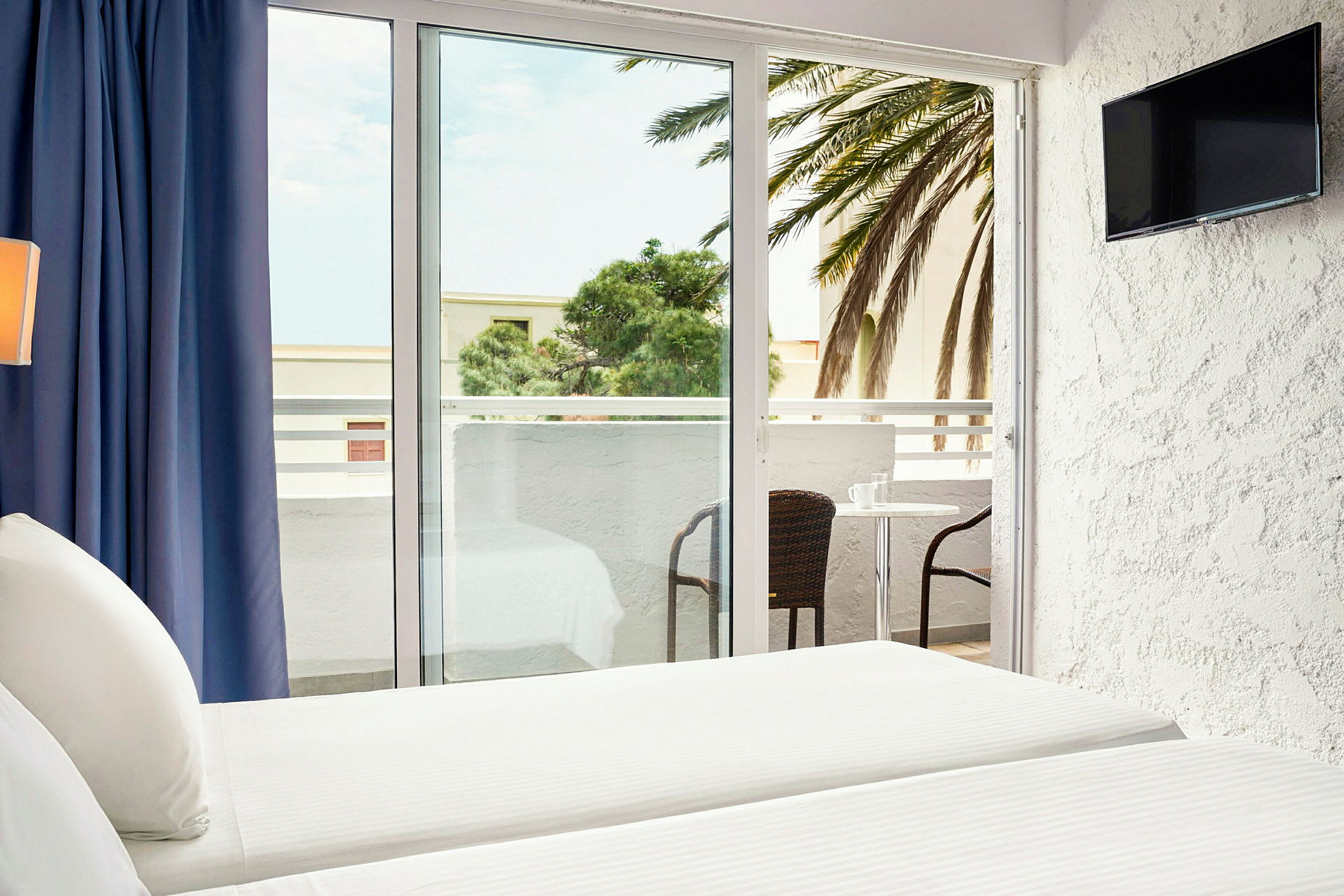 Ett hotellrum med vita sängkläder och en balkong med bord och stolar. Utsikten visar en palm och byggnader. (AI-genererad text)