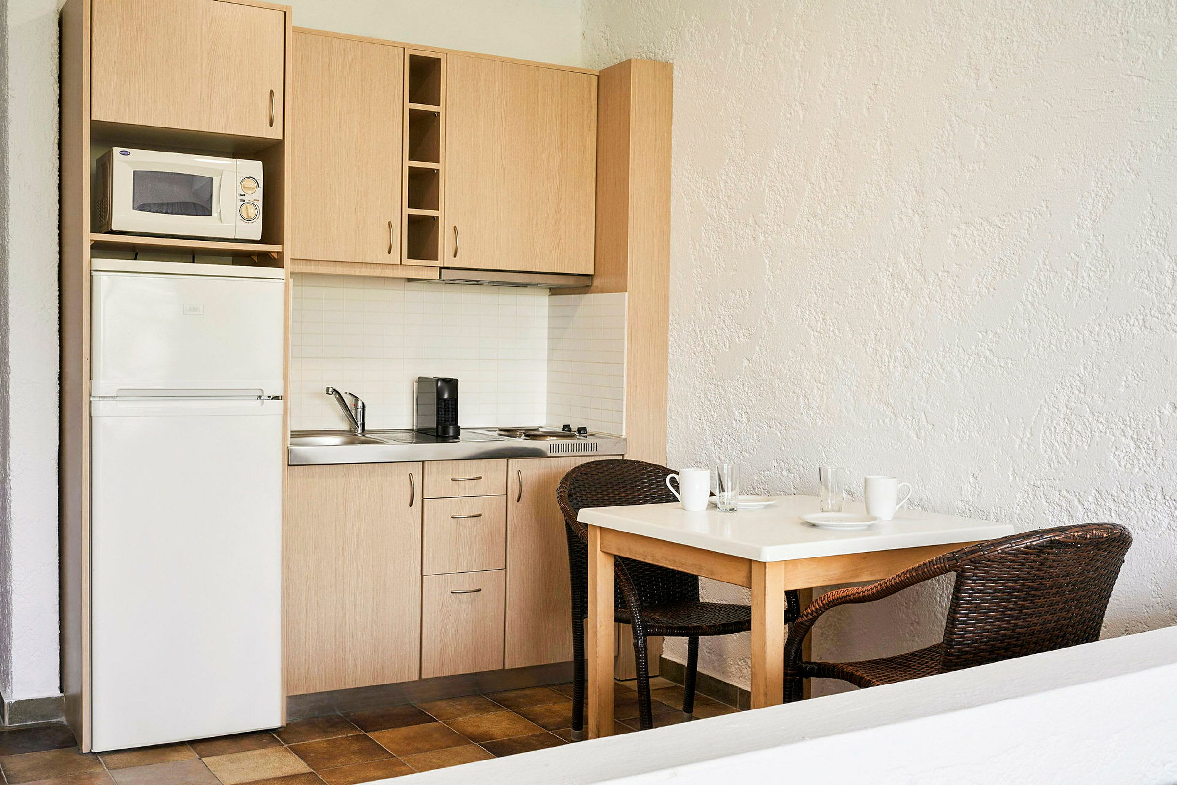Ett kompakt kök med träskåp, en mikrovågsugn, ett kylskåp och en kaffemaskin. Framför köket står ett bord med två rottingstolar. (AI-genererad text)