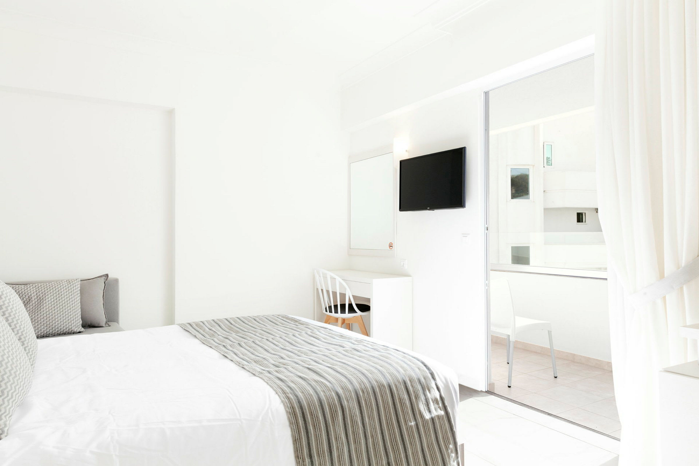 Interiören av ett ljust hotellrum med en säng och skrivbord bredvid en vit stol. En TV är monterad på väggen och glasdörrar leder till en balkong. (AI-genererad text)