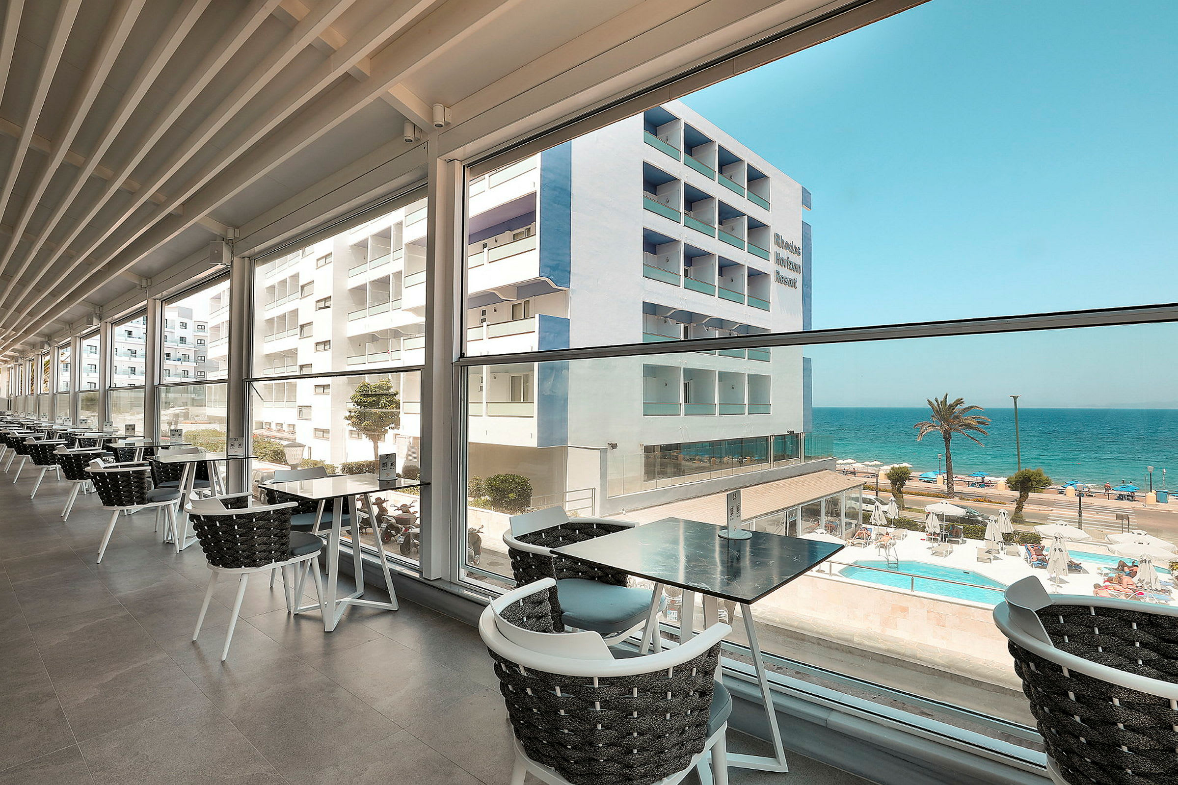 Utsikt från en restaurang med glasväggar mot en byggnad, en pool och havet. (AI-genererad text)