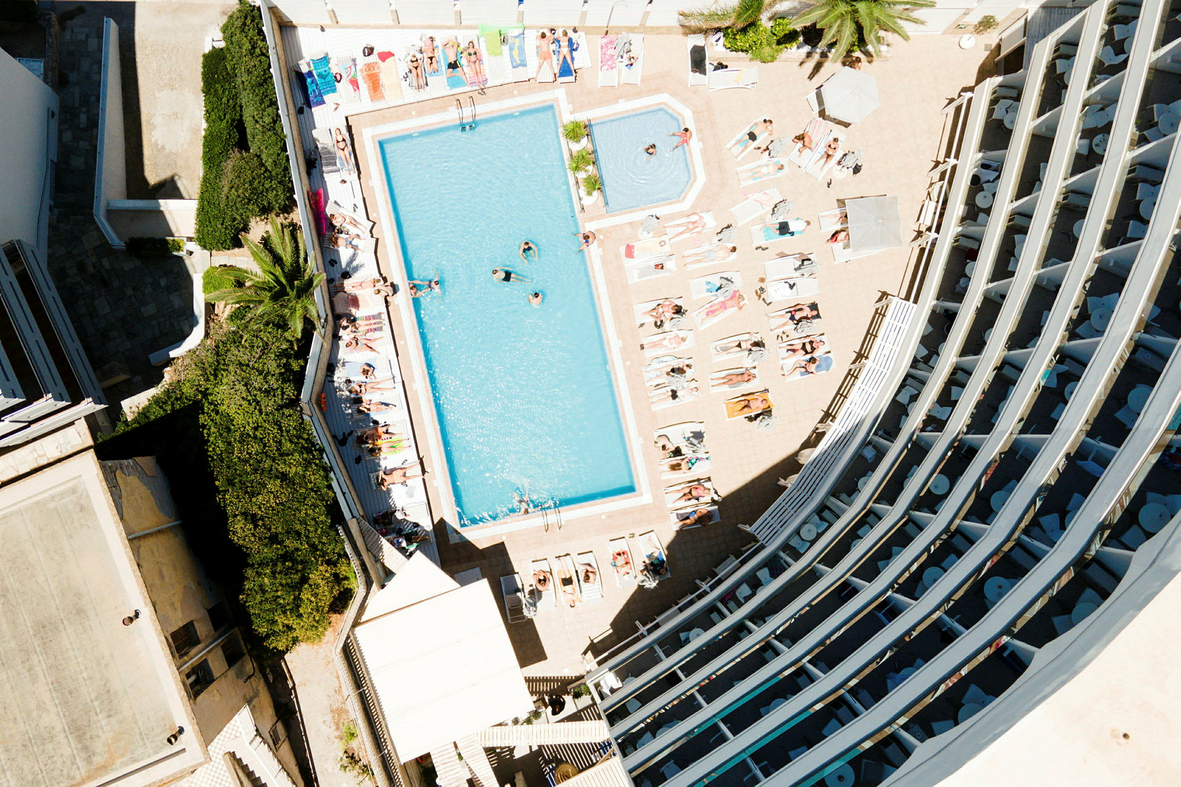 Flygfoto av en poolområde vid ett hotell, med solstolar, människor och omgivande byggnader. (AI-genererad text)