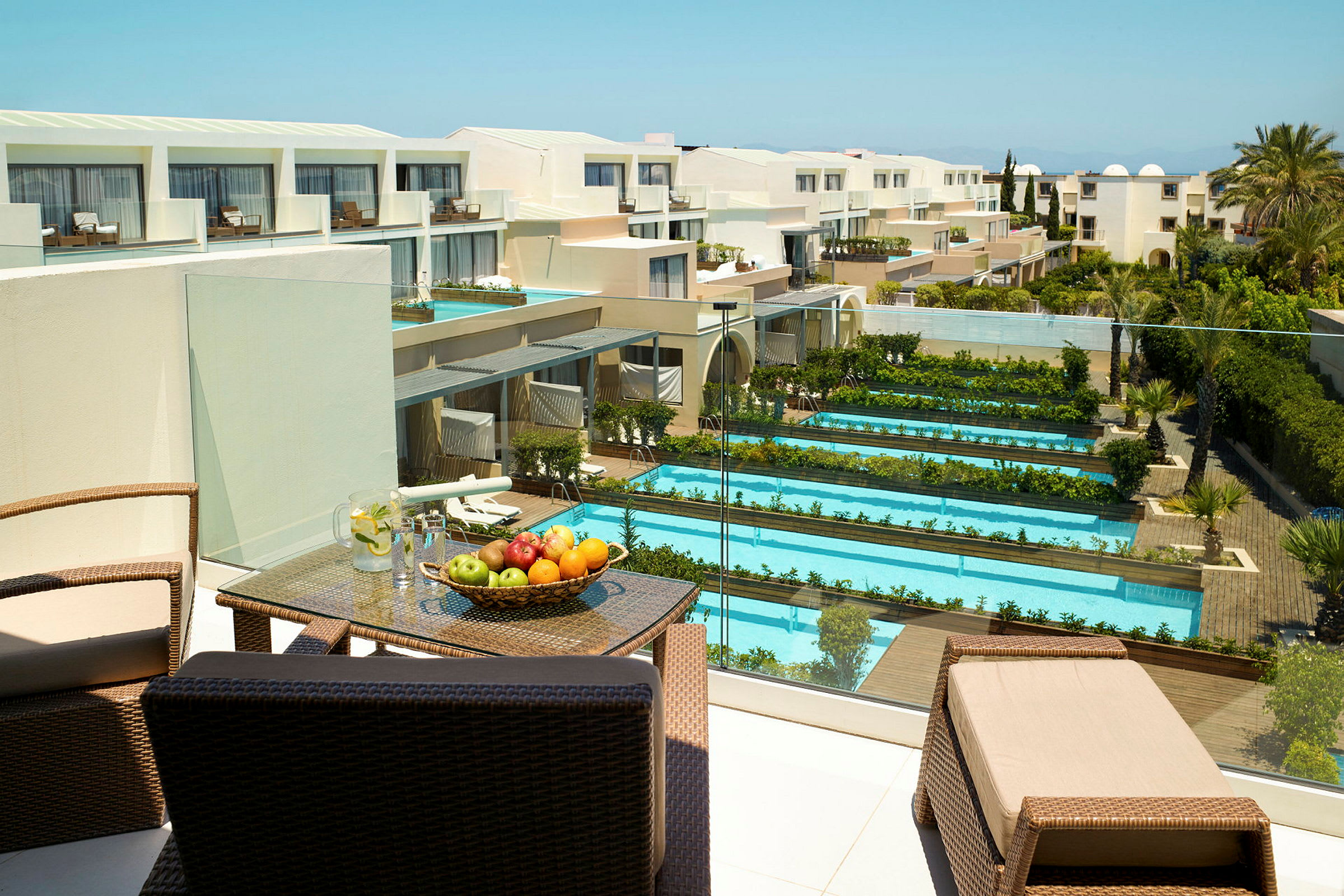 Hotellområde med flera pooler och grönområden, samt en terrass med möbler och frukt. Utsikt över en modern byggnad. (AI-genererad text)