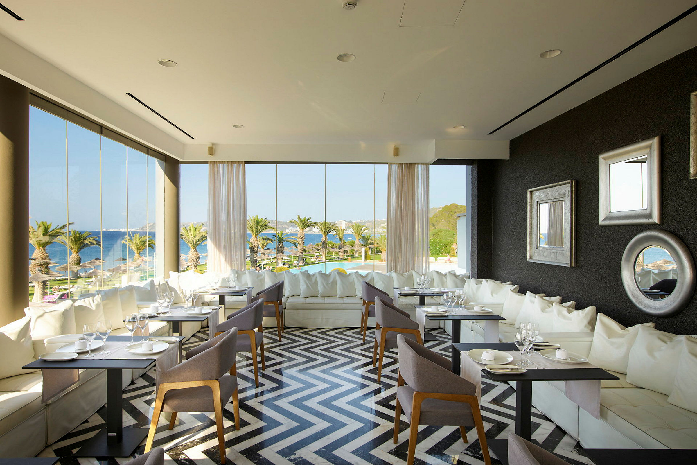 Interiör av en restaurang med stora fönster, moderna möbler och utsikt över palmer och havet. (AI-genererad text)