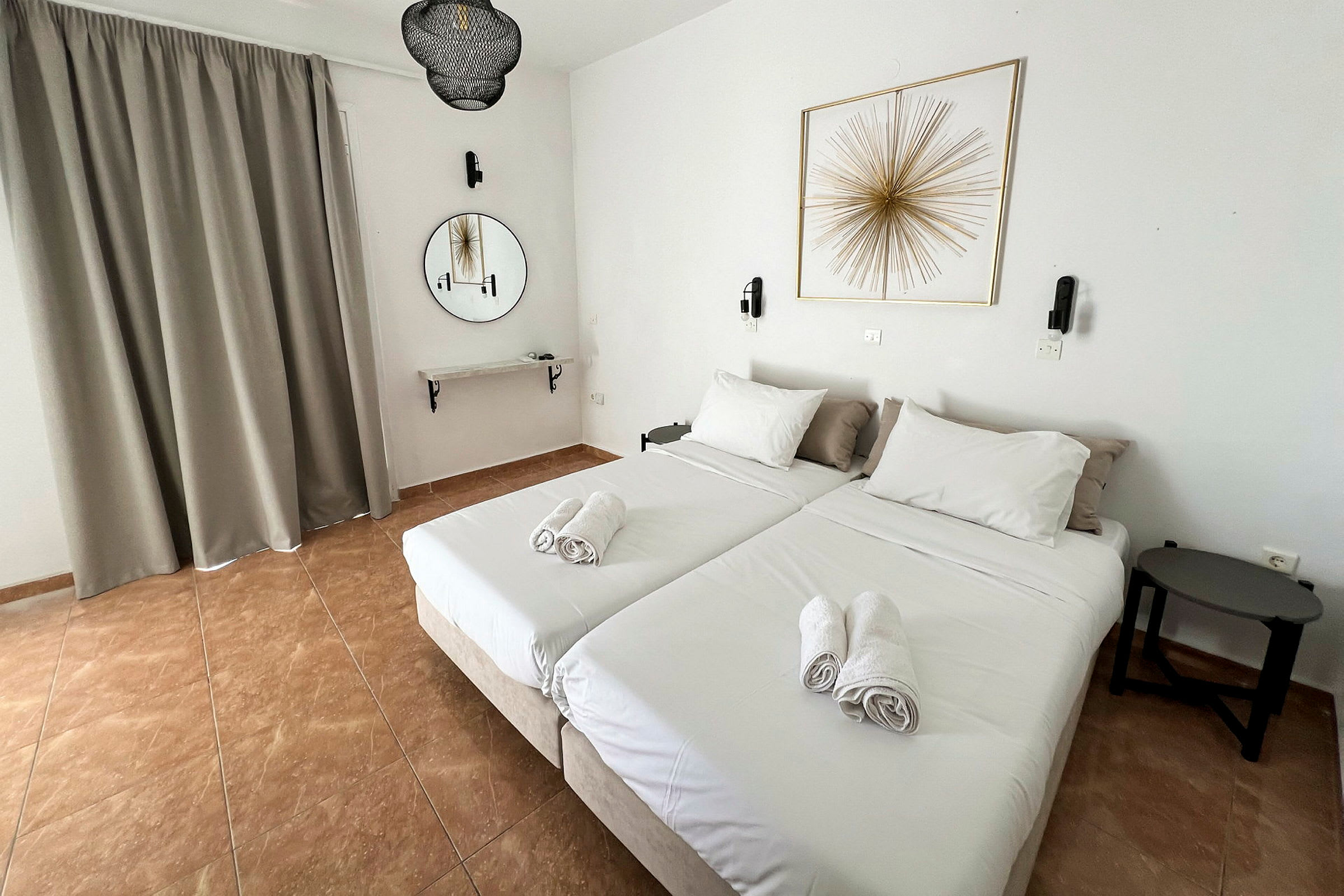 Två enkelsängar med vita sängkläder och vikta handdukar ovanpå står i ett hotellrum. Ett runt spegel och en väggdekoration syns på väggen. (AI-genererad text)