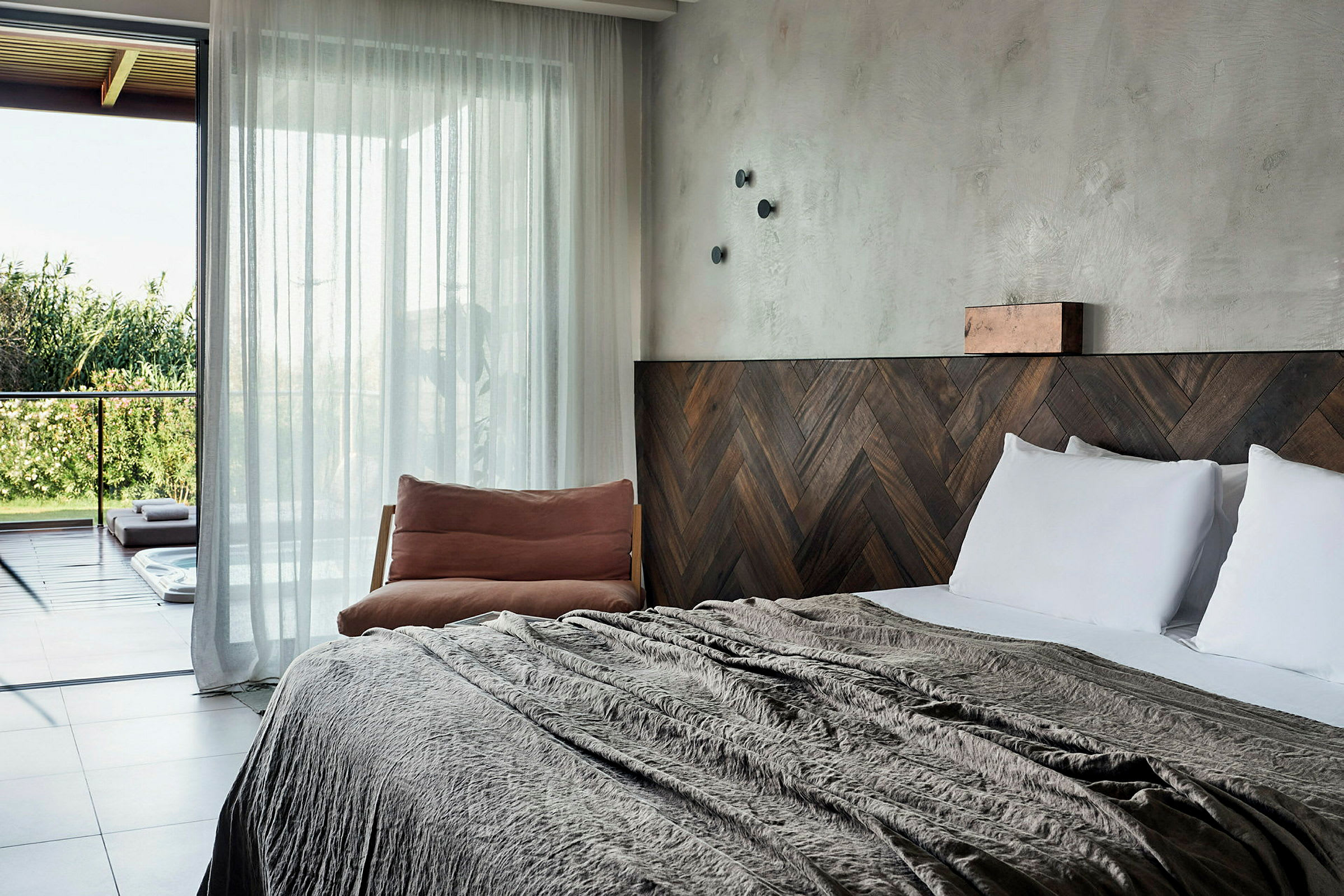 Ett hotellrum med en stor säng, trädekorationer och en stol vid sidan, samt glasdörrar som leder ut till en terrass med utsikt över grönska. (AI-genererad text)