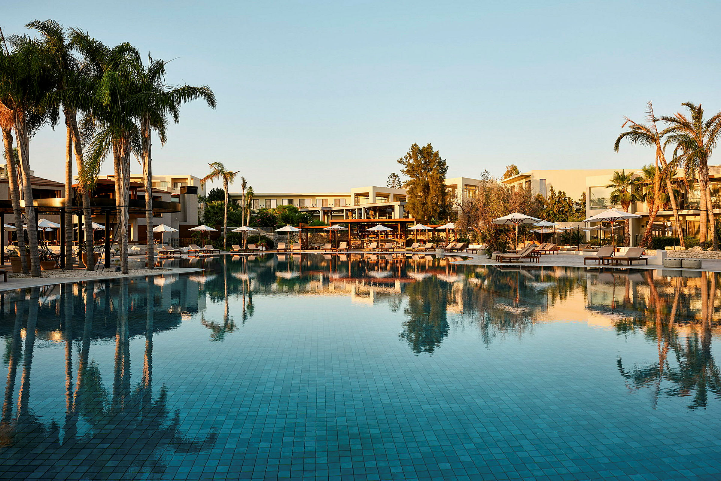 Stort poolområde med palmer och solstolar med parasoll. I bakgrunden syns hotellbyggnader. (AI-genererad text)