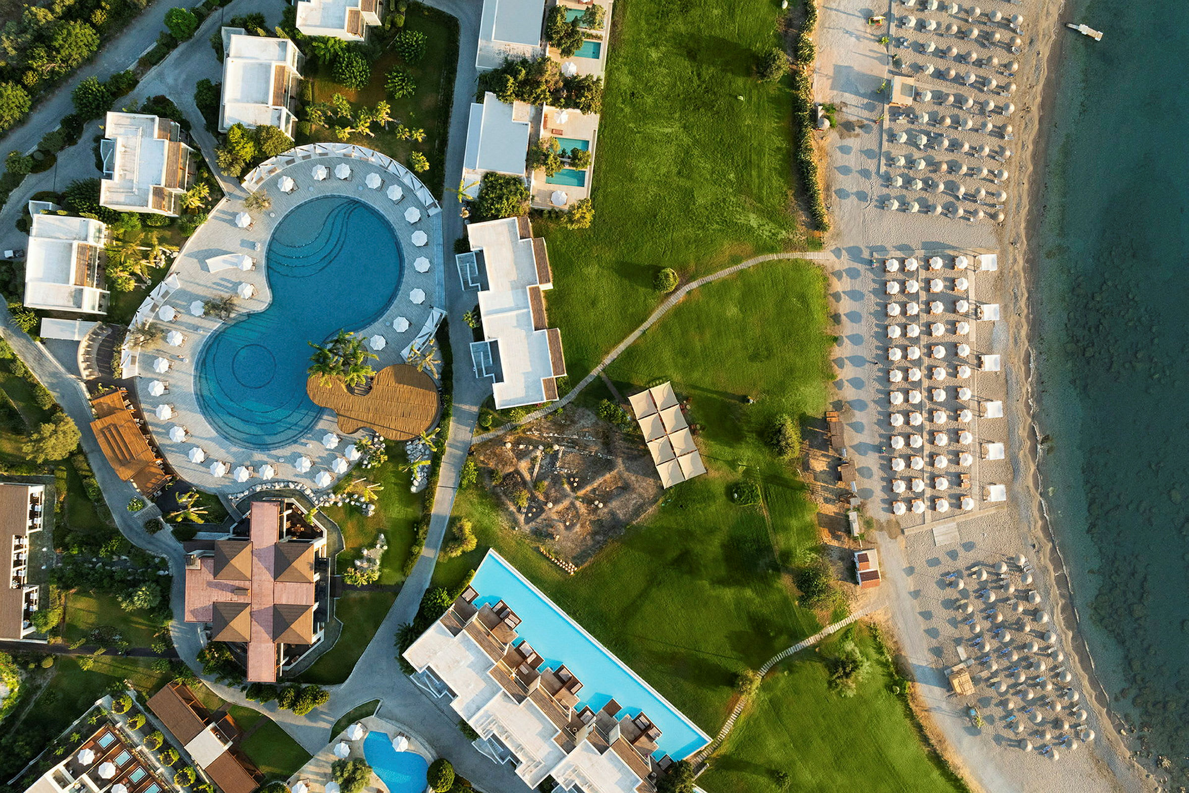 Flygfoto över en semesteranläggning med pool, grönytor och strand med parasoll. (AI-genererad text)