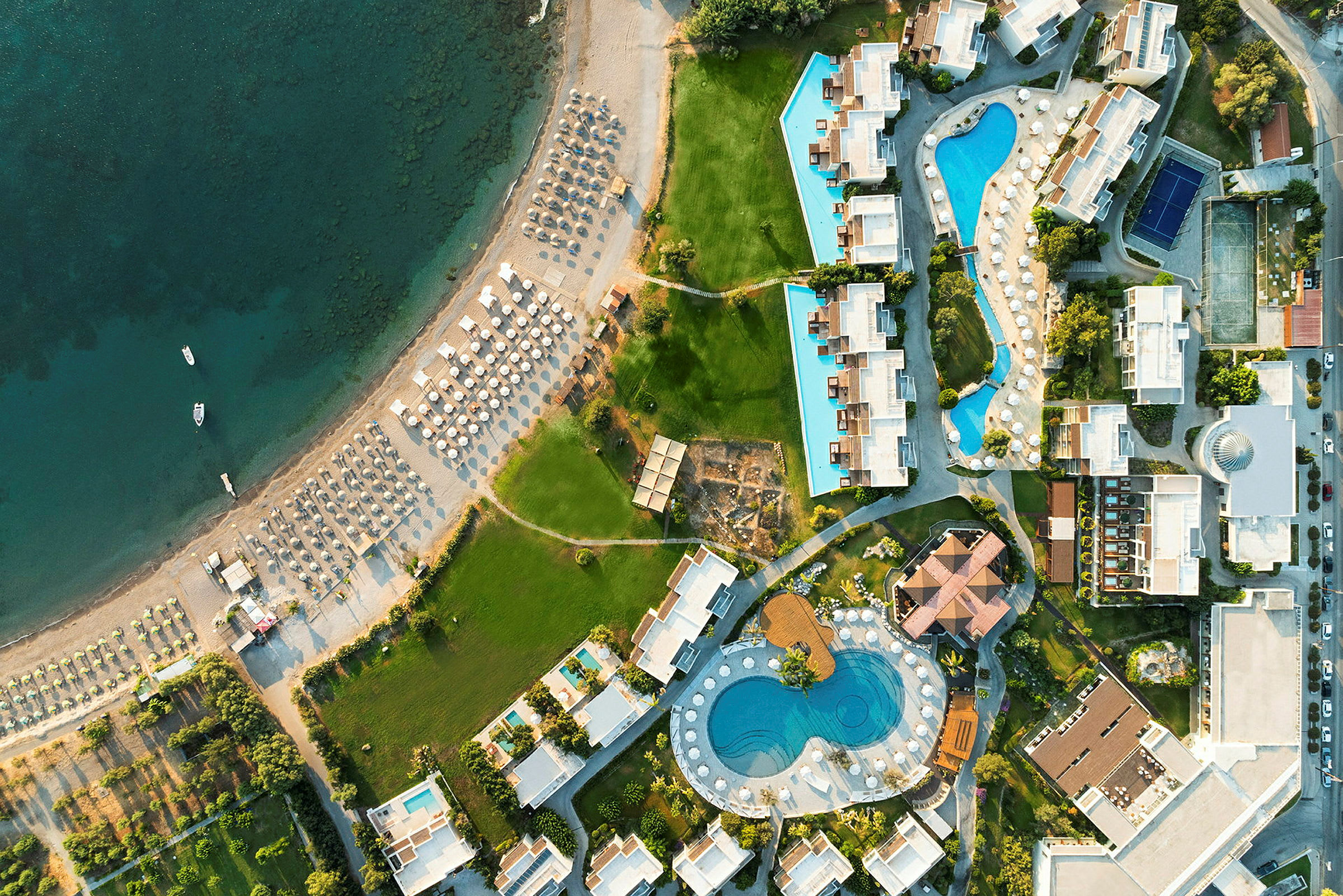 Flygbild av en badort med byggnader, swimmingpooler och en strand med solstolar. (AI-genererad text)