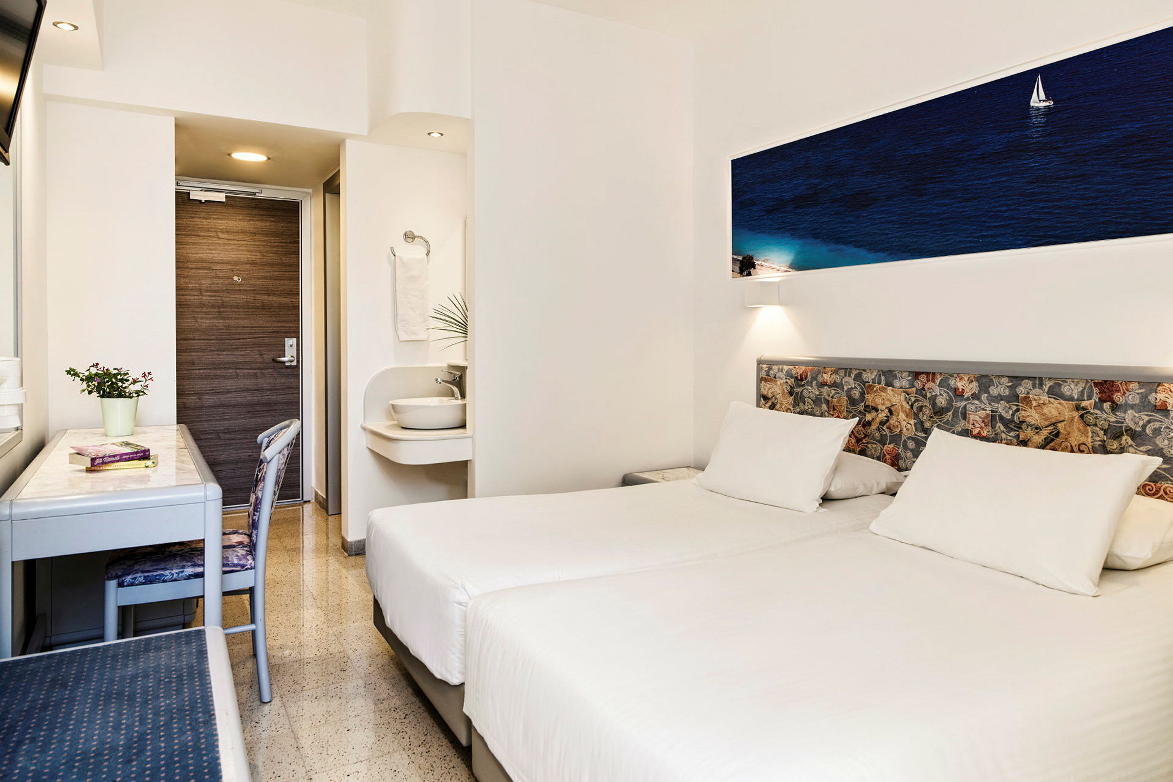 Ett hotellrum med två vita enkelsängar, ett skrivbord, en stol och en liten handfat utanför badrummet. (AI-genererad text)