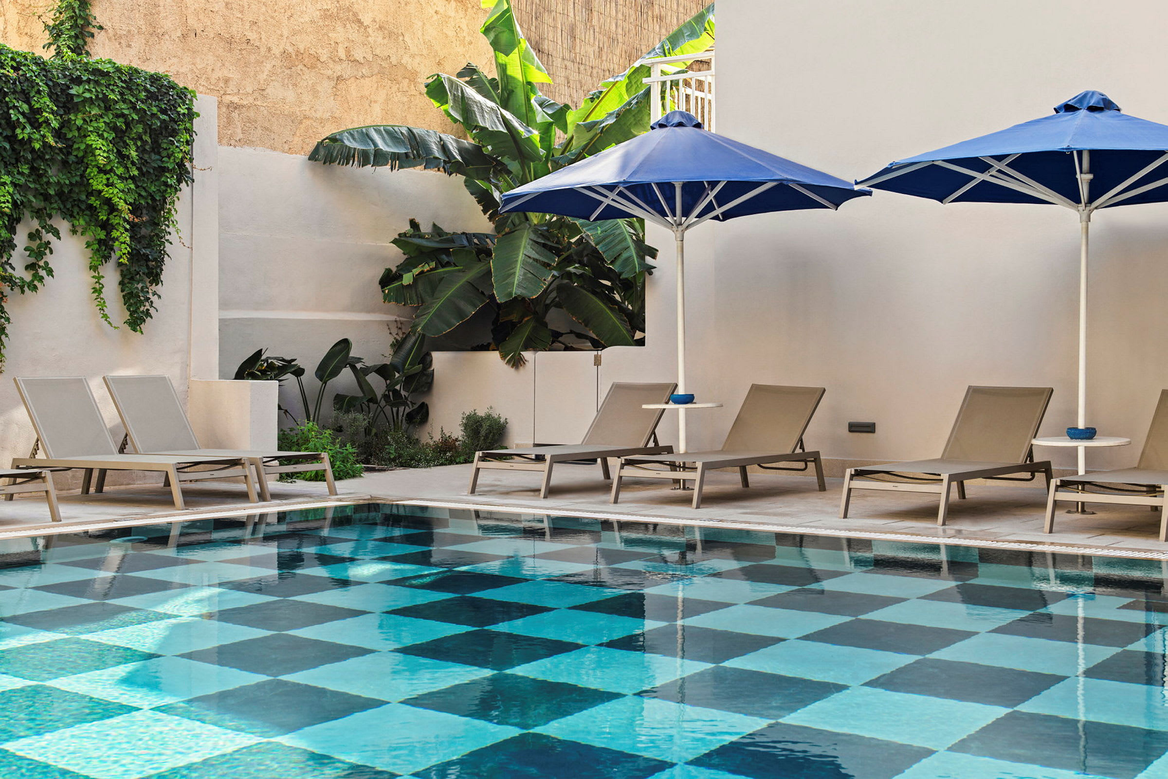 Rektangulär pool med ett schackmönstrat golv omgiven av solstolar och parasoller. (AI-genererad text)