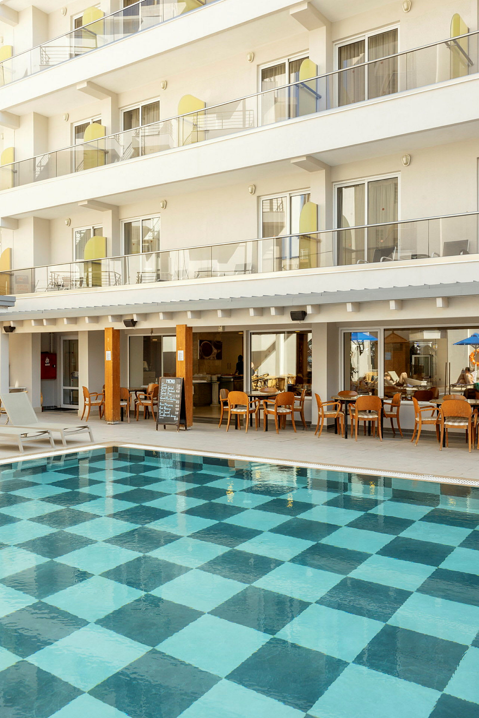 Utomhuspool med rutigt bottenmönster framför hotellbyggnad med balkonger och restaurang. (AI-genererad text)