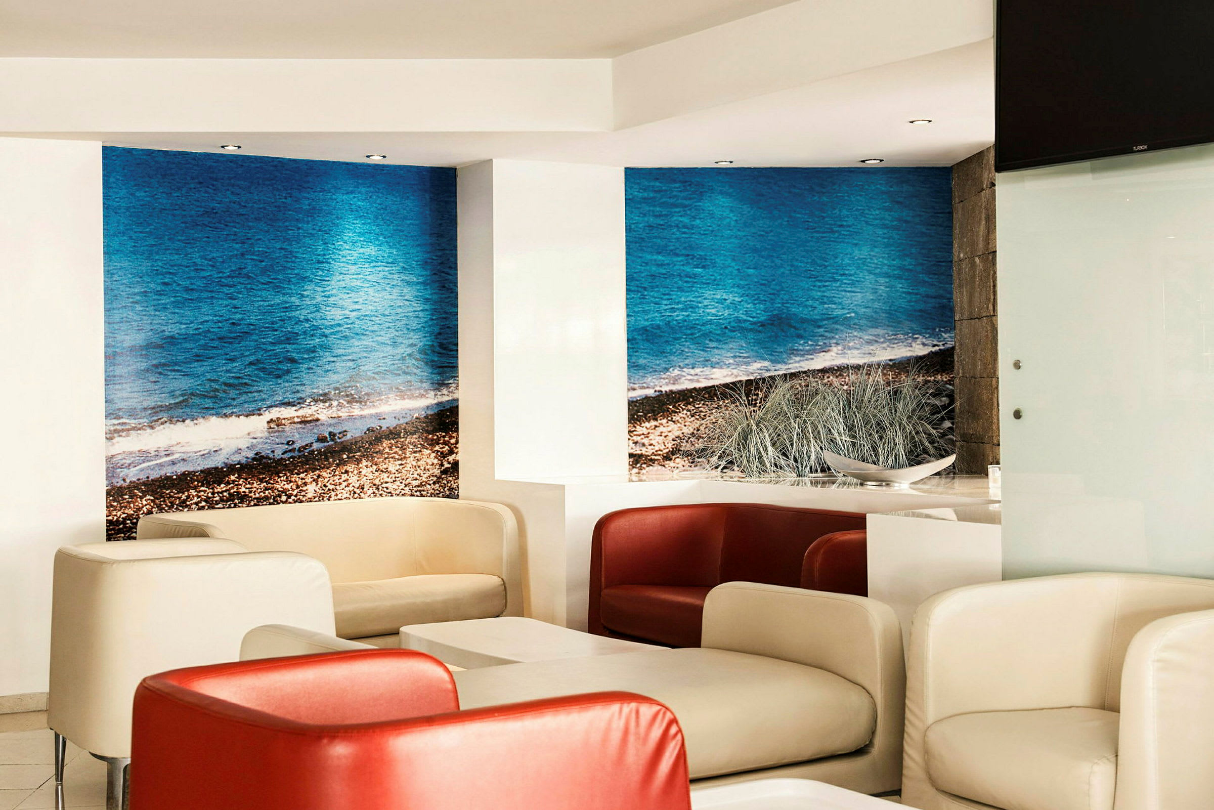 En modern lounge med bekväma stolar i vitt och rött samt bord. Väggarna har en fototapet med motiv av en strand med blått hav. (AI-genererad text)