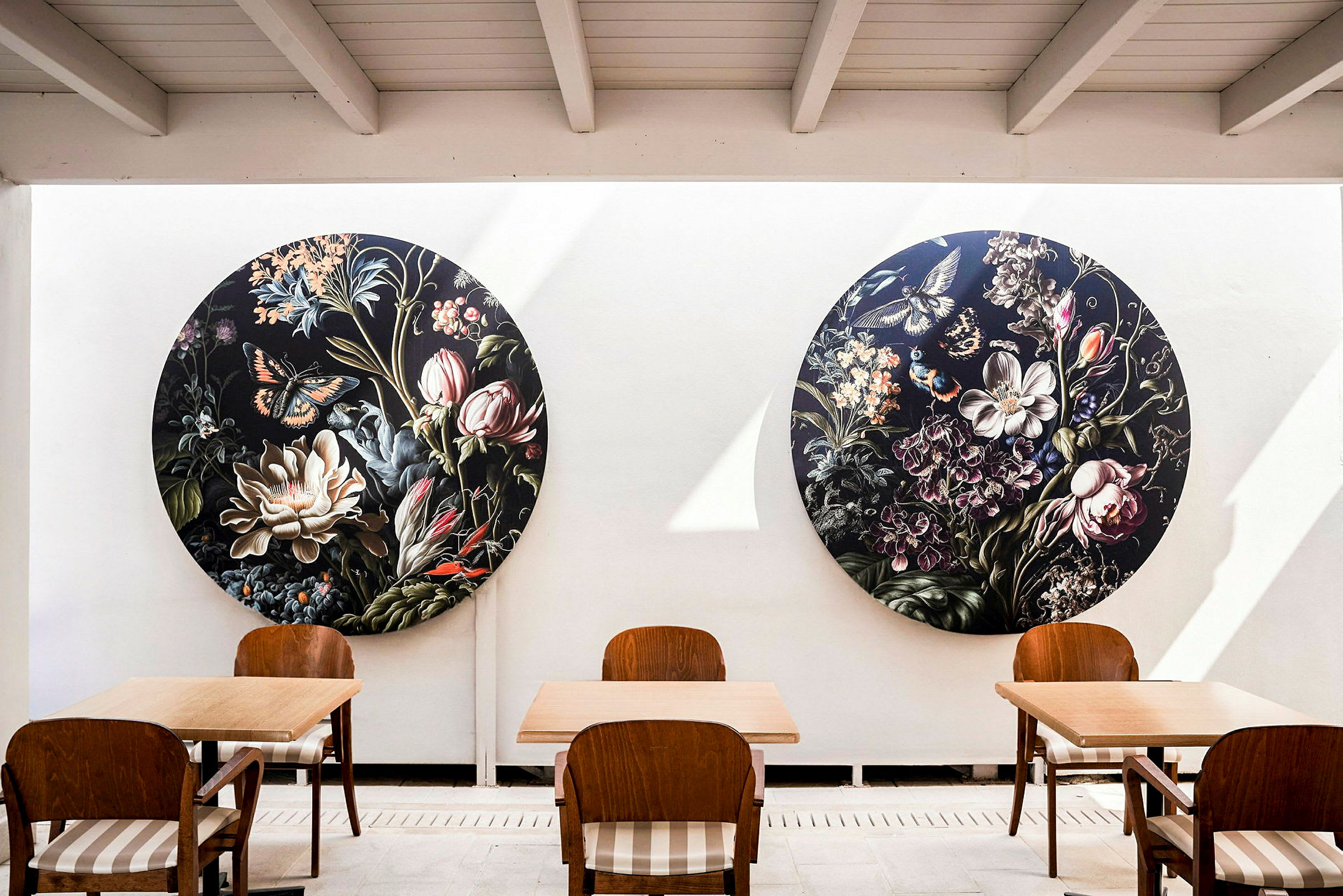 Bild som visar en stilfull interiör med två runda tavlor föreställande blommor och fjärilar på en vit vägg. Framför väggen står trämöbler med stolar och bord. (AI-genererad text)