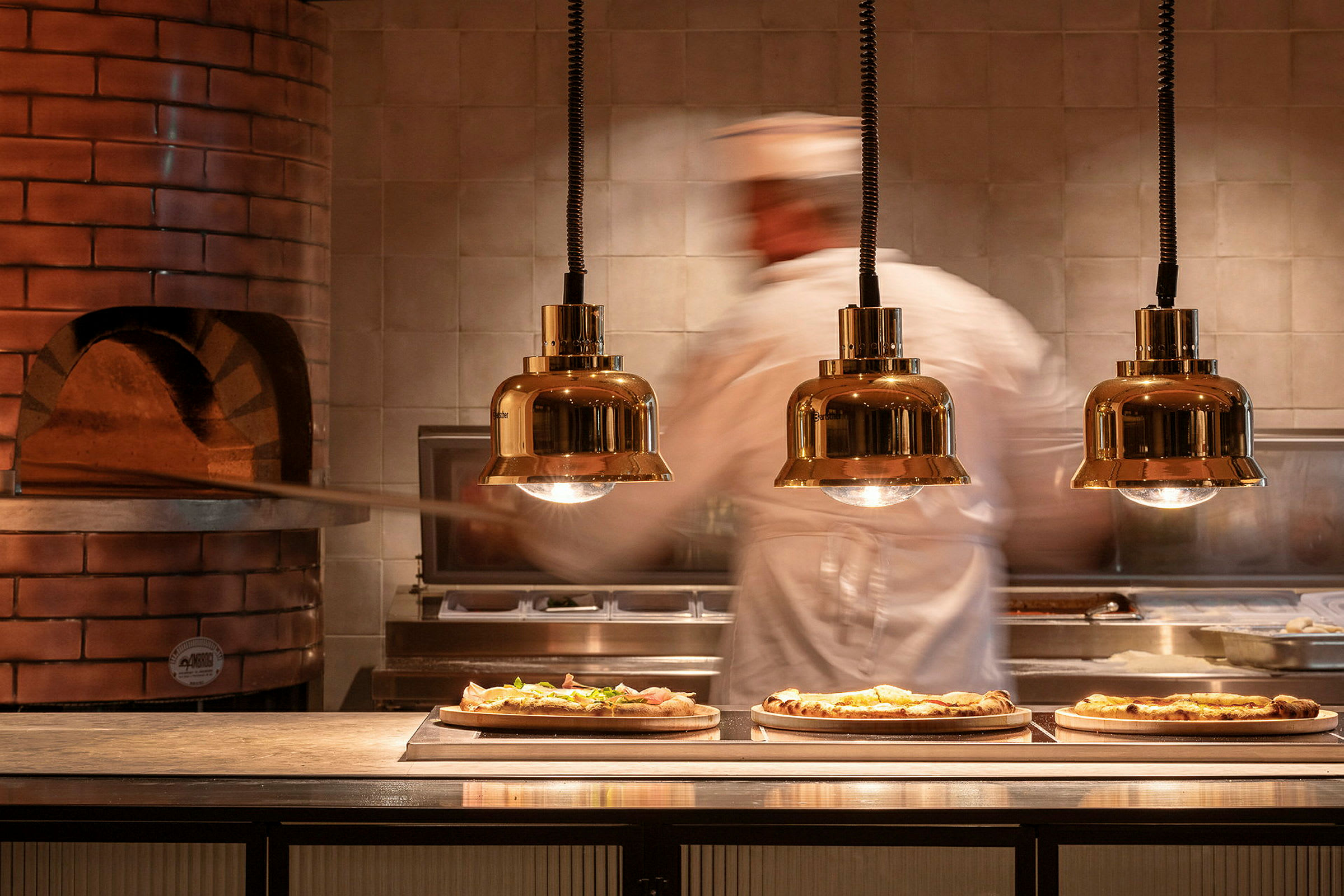 En pizzabagare arbetar vid en ugn där tre färdiga pizzor ligger på serveringsbordet. (AI-genererad text)